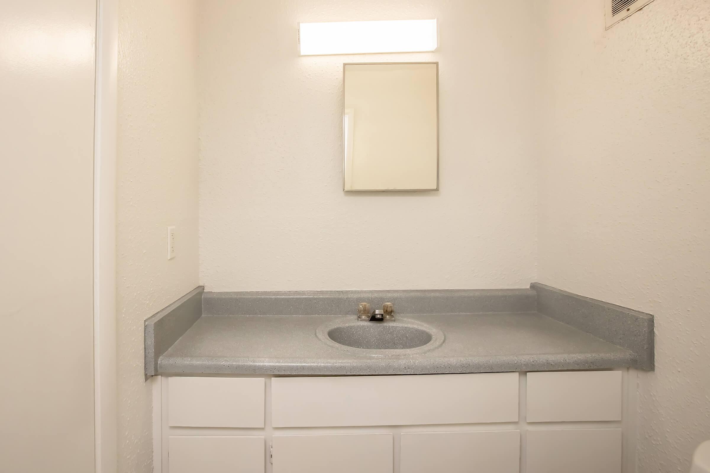 A simple bathroom with a light-colored wall and a gray countertop. There is a circular sink with faucet fixtures and a mirror above it. The cabinetry below the countertop is white with a minimalist design, and the lighting is provided by a wall-mounted fixture above the mirror.