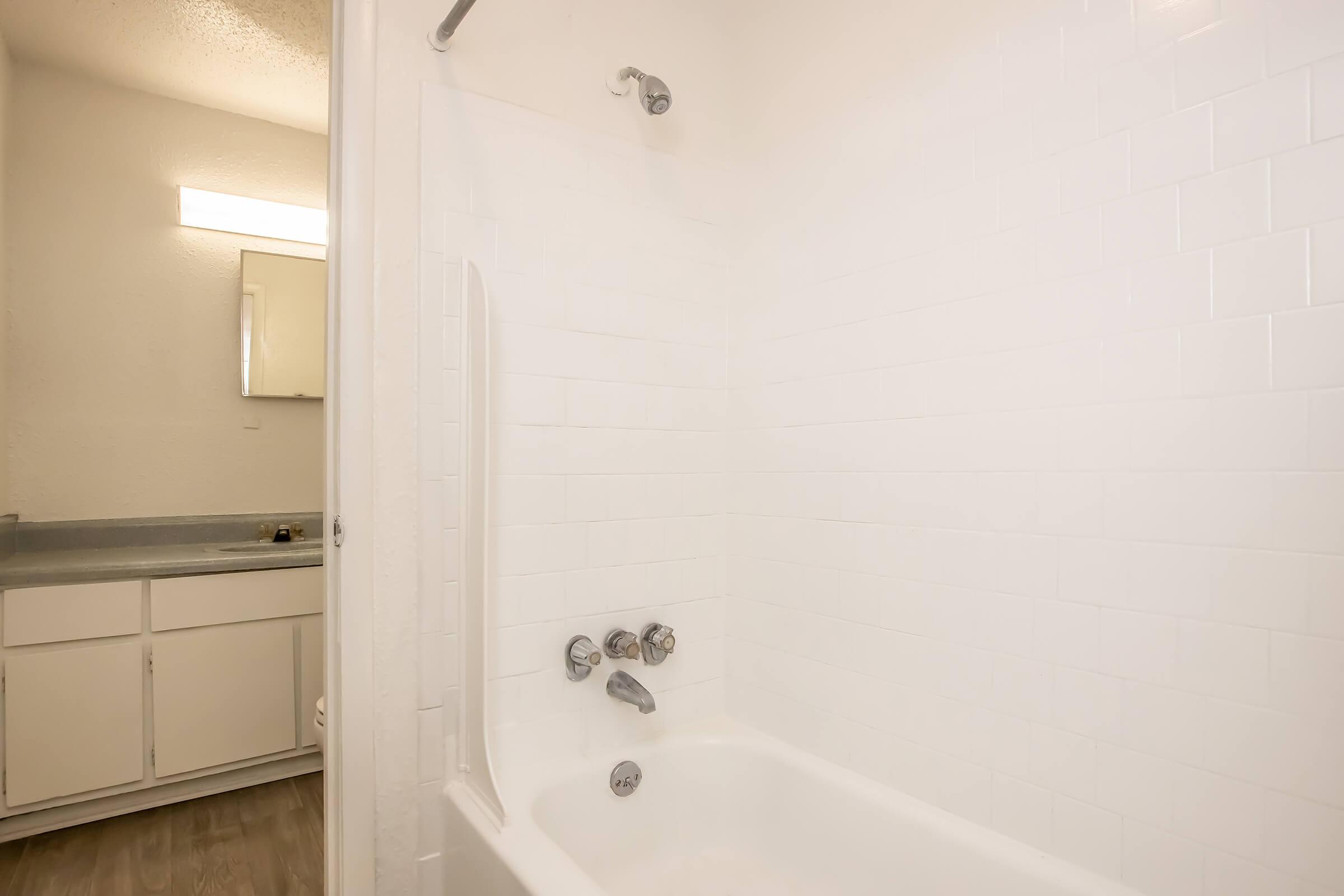 A clean and modern bathroom featuring a white tiled bathtub with chrome fixtures. The bathtub is situated against a light-colored wall, and in the background, a glimpse of a vanity and mirror can be seen, suggesting a well-maintained and bright space.