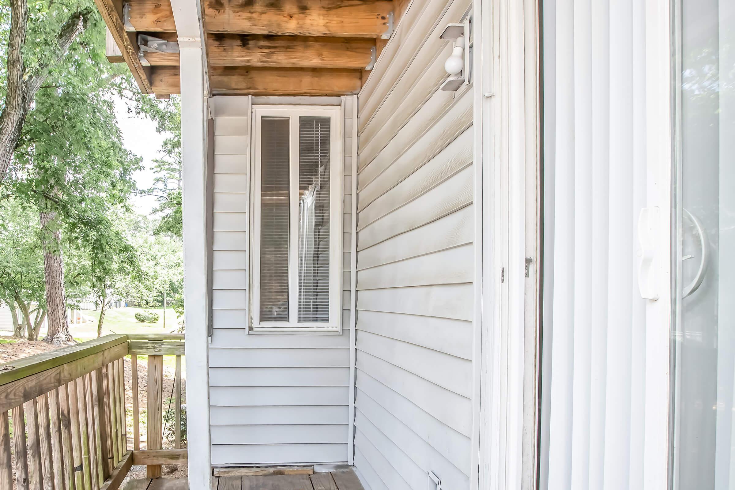 A corner of a house showing a wooden deck, with a small window and a white door on the side. The siding is light-colored, and there are trees visible in the background, indicating an outdoor setting.