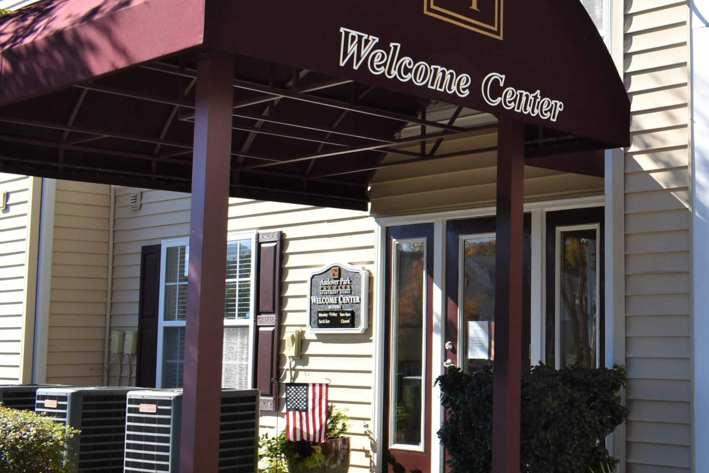 A photo of a building entrance featuring a maroon awning with the text "Welcome Center." The entrance includes large windows, an American flag displayed on the side, and a decorative sign next to the door, all set against a beige siding backdrop.