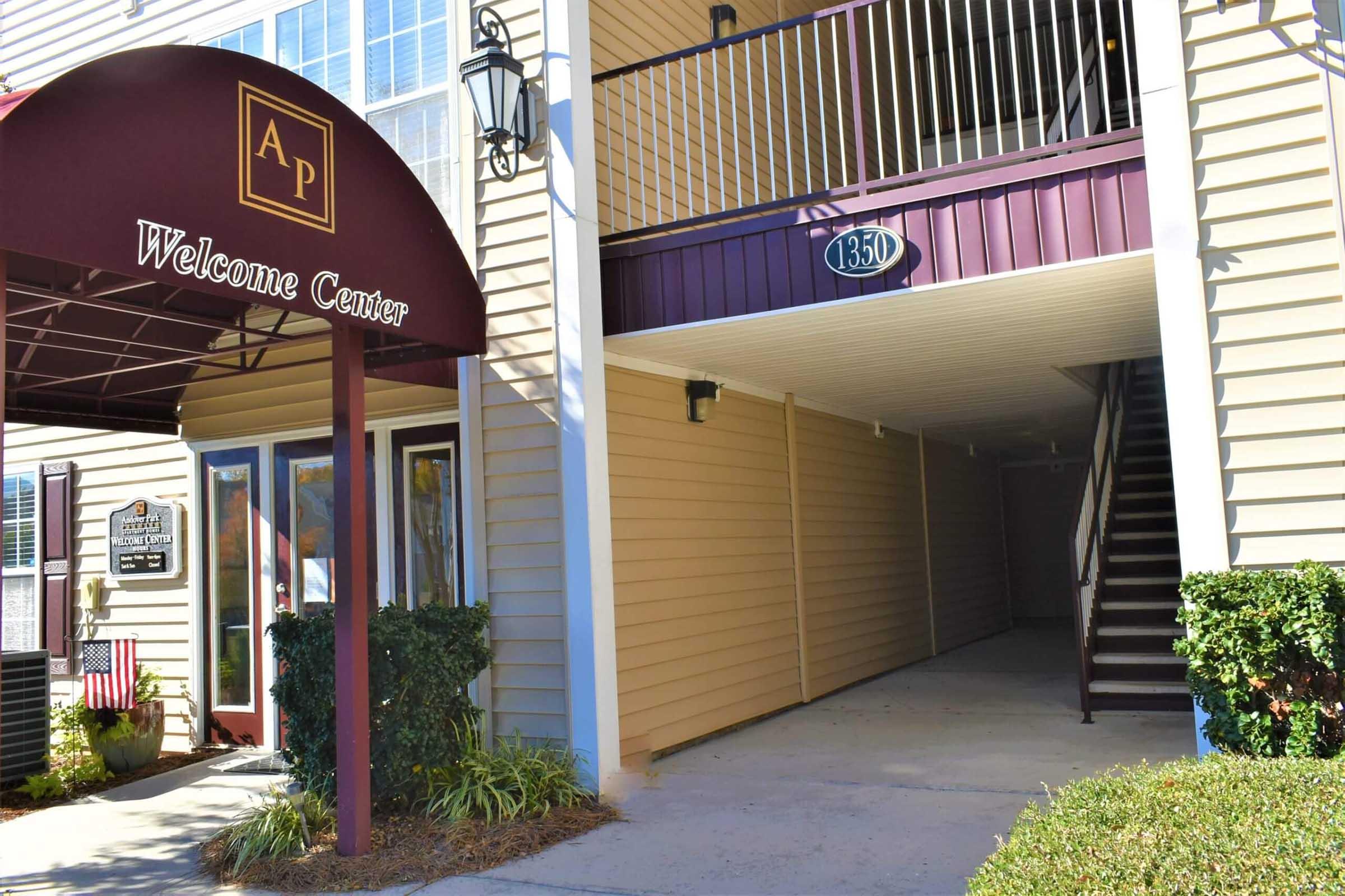 Entrance to a welcome center with a maroon awning displaying "Welcome Center" and the initials "AP." The building features beige siding and a staircase visible in the background, alongside a sign indicating the unit number 1350. Lush greenery lines the entrance pathway.