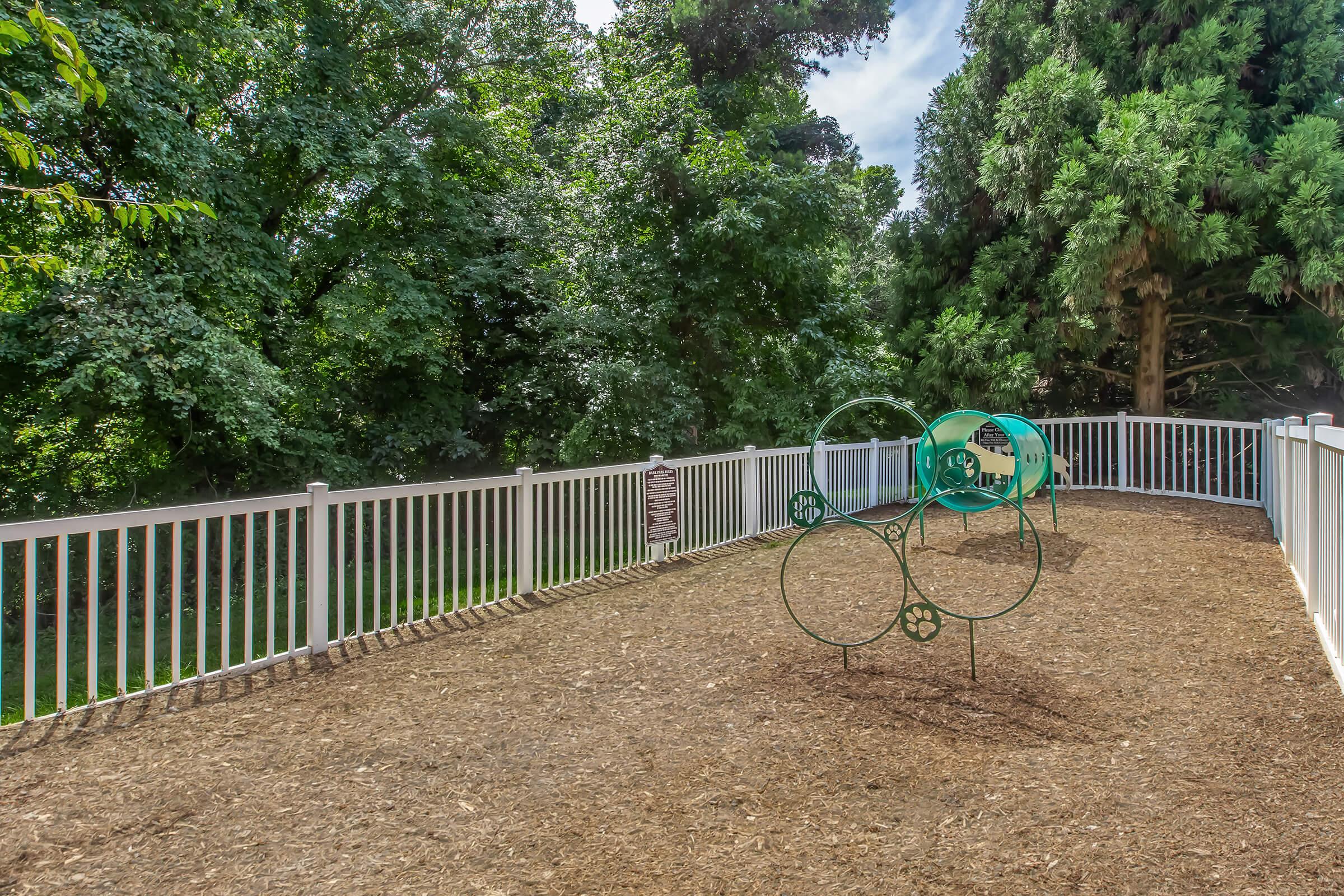 A playground area featuring green metal equipment for climbing and play, surrounded by a white fence. The ground is covered with wood mulch, and tall trees provide a lush green backdrop under a clear blue sky.