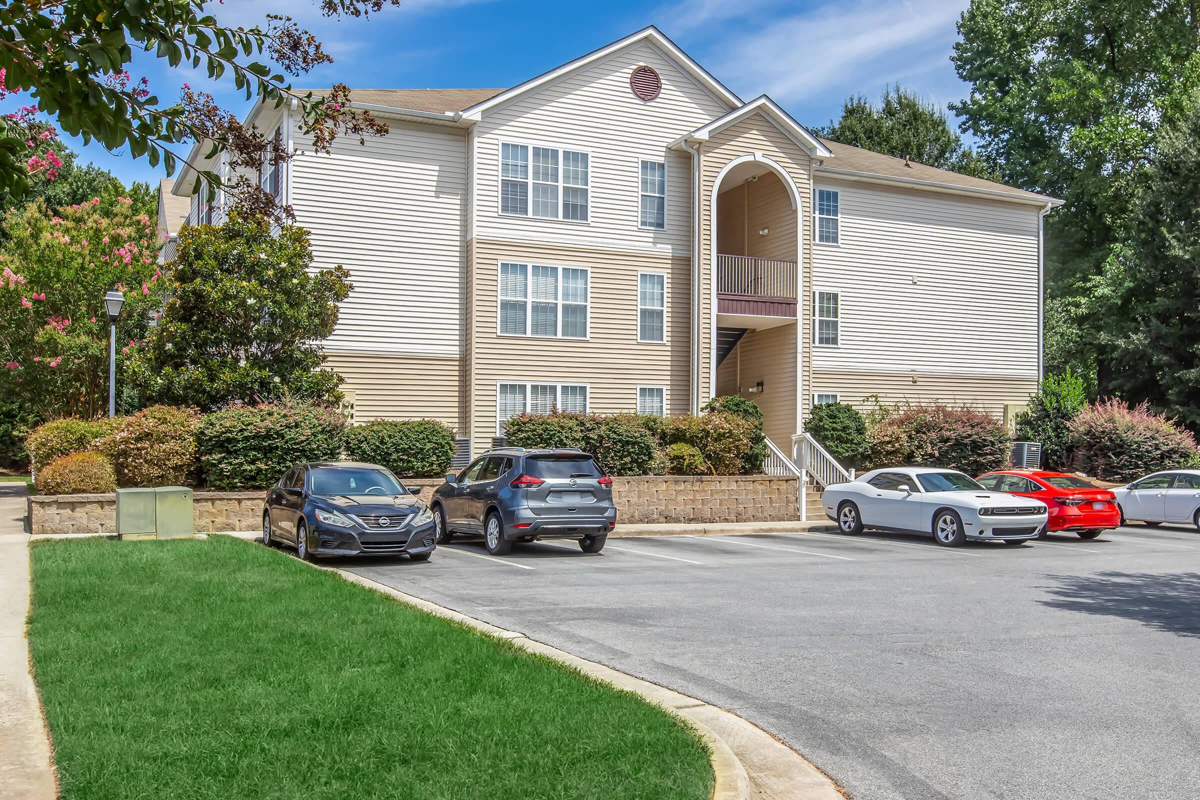 A three-story apartment building with a beige exterior and white trim, surrounded by green shrubbery and trees. Several cars are parked in the vacant lot in front of the building, including a gray SUV, a red sports car, and a white sedan. The sky is clear with a few clouds.