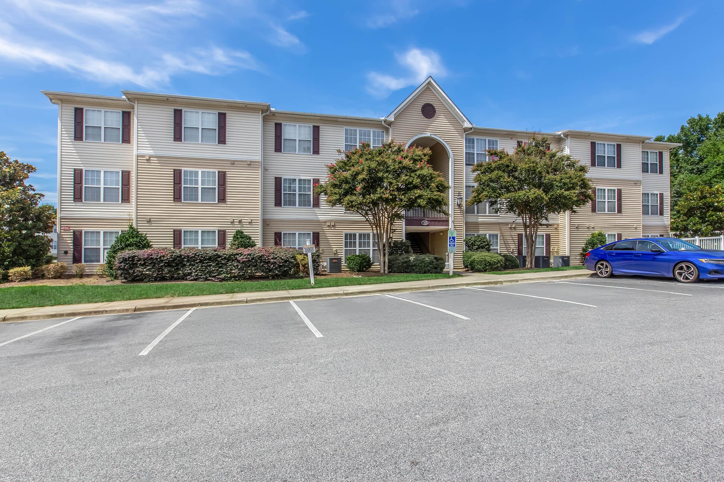 Exterior view of a multi-story apartment building with a beige facade and multiple windows, surrounded by neatly trimmed shrubs and small trees. The building features a central archway entrance and is set back from a parking lot with empty spaces. The sky is blue with a few clouds.