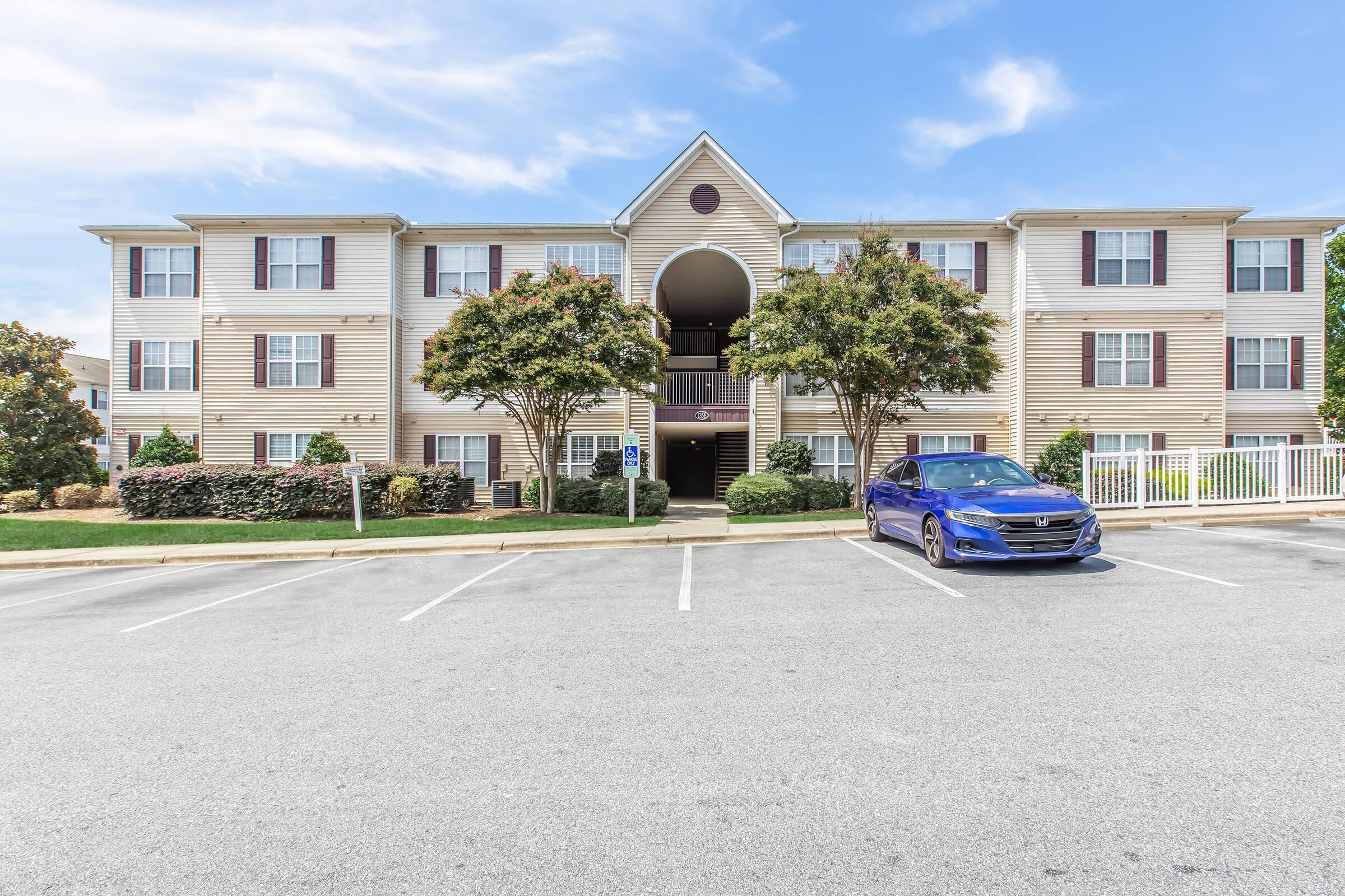 A two-story residential building with a light-colored exterior, featuring multiple windows and a central archway. In the foreground, there is a gray parking lot with several empty spaces and a blue car parked at the far right. Well-maintained landscaping surrounds the building, with shrubs and trees.