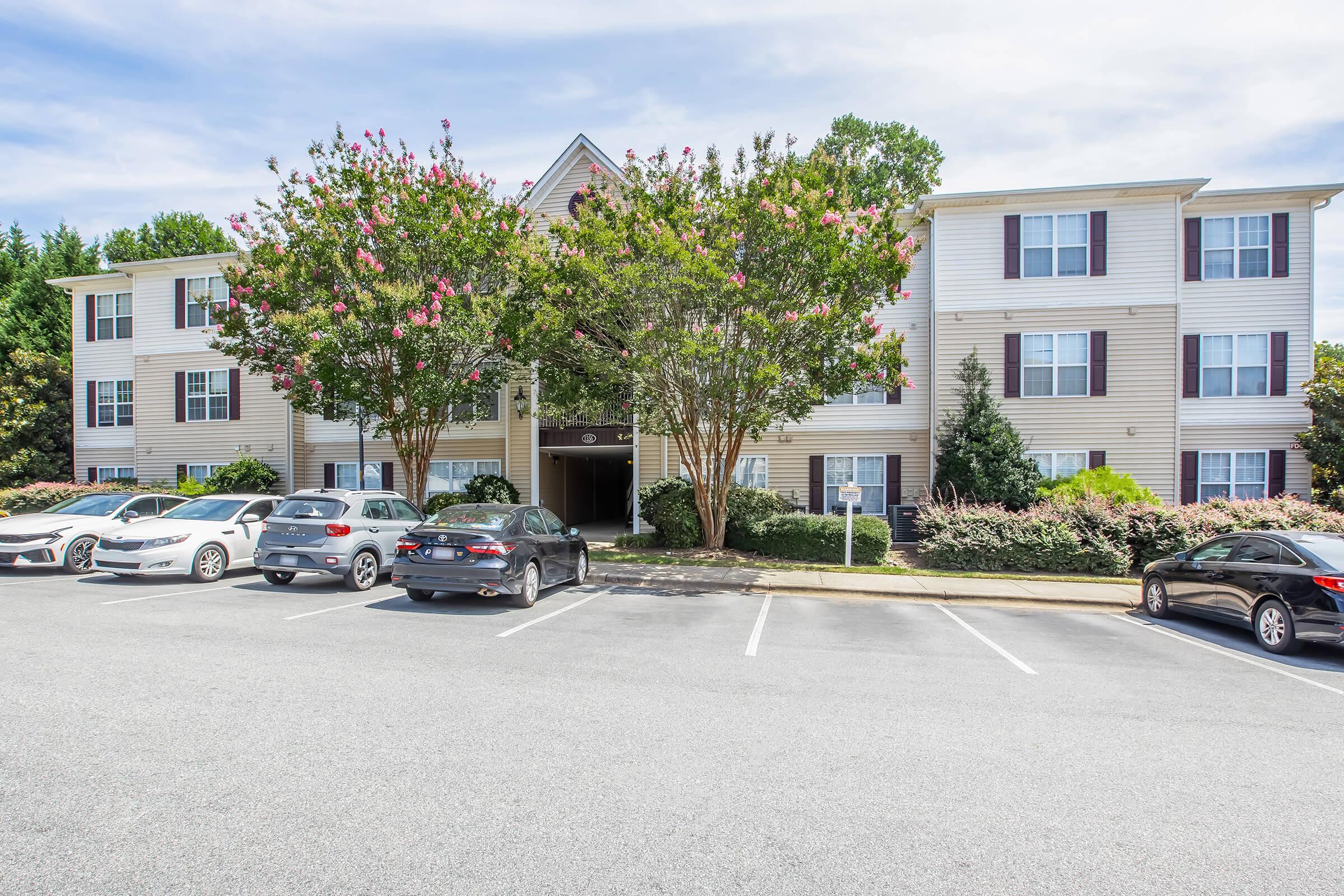A three-story residential building with multiple windows, surrounded by blooming trees and landscaping. Several parked cars are visible in the foreground, along with a paved parking lot. The sky is partly cloudy, creating a bright and inviting atmosphere.