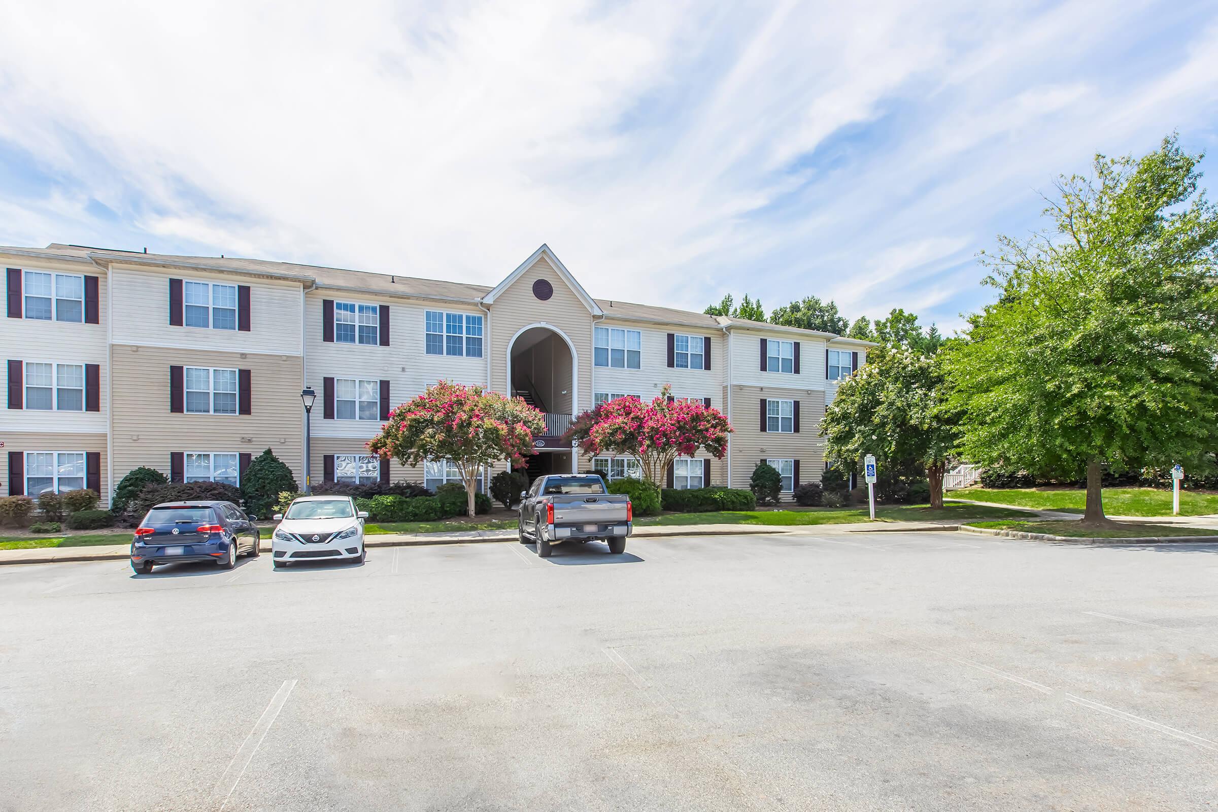 Exterior view of a three-story residential building with beige siding, featuring balconies and blooming pink flowers. The parking lot in front has several parked cars, and there are landscaped trees and grass areas surrounding the entrance. The sky is partly cloudy.