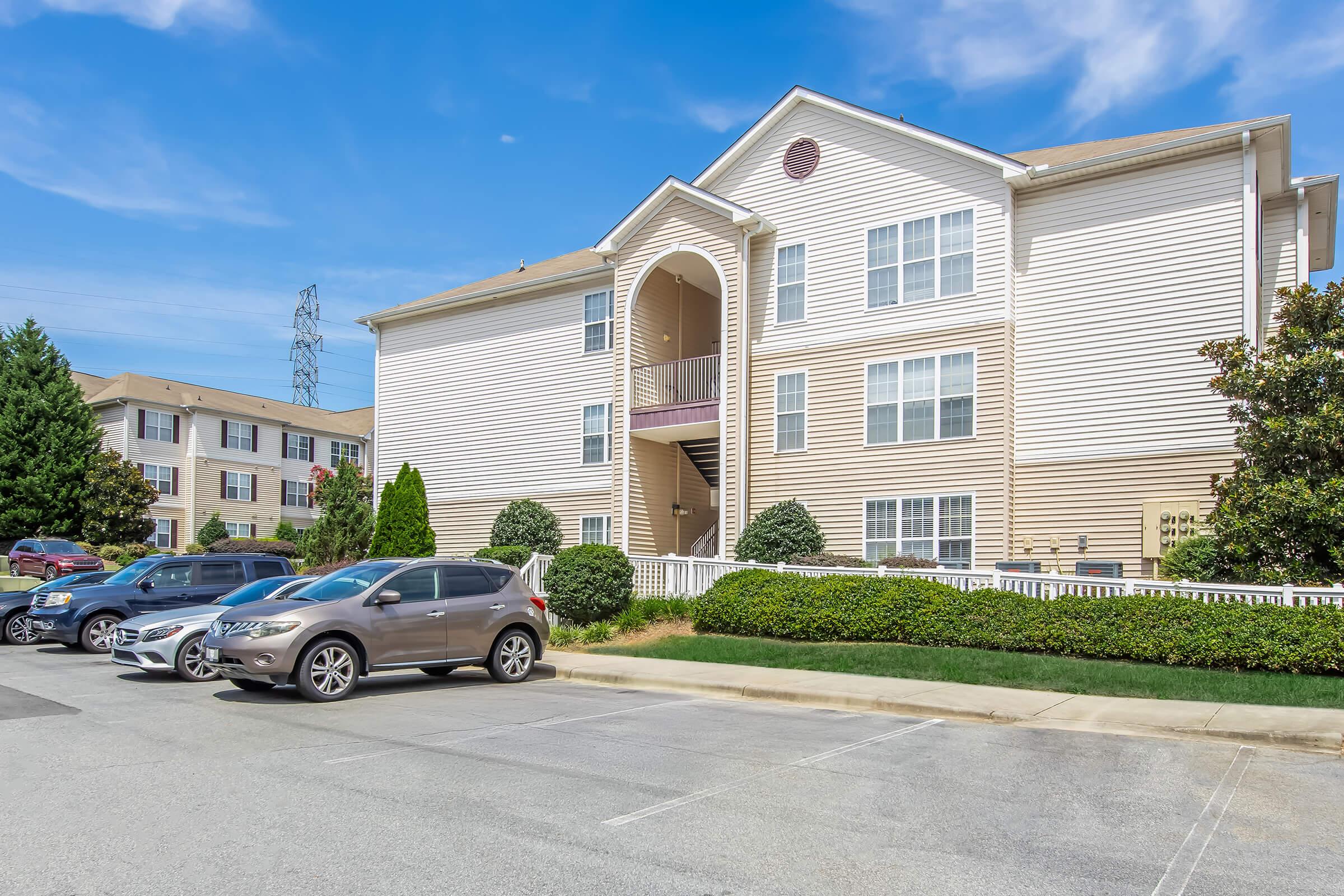 A three-story apartment building with light-colored siding and multiple windows. In the foreground, parked cars line a driveway, and well-maintained shrubs and small trees are visible. The sky is clear with a few clouds, and another similar building can be seen in the background.