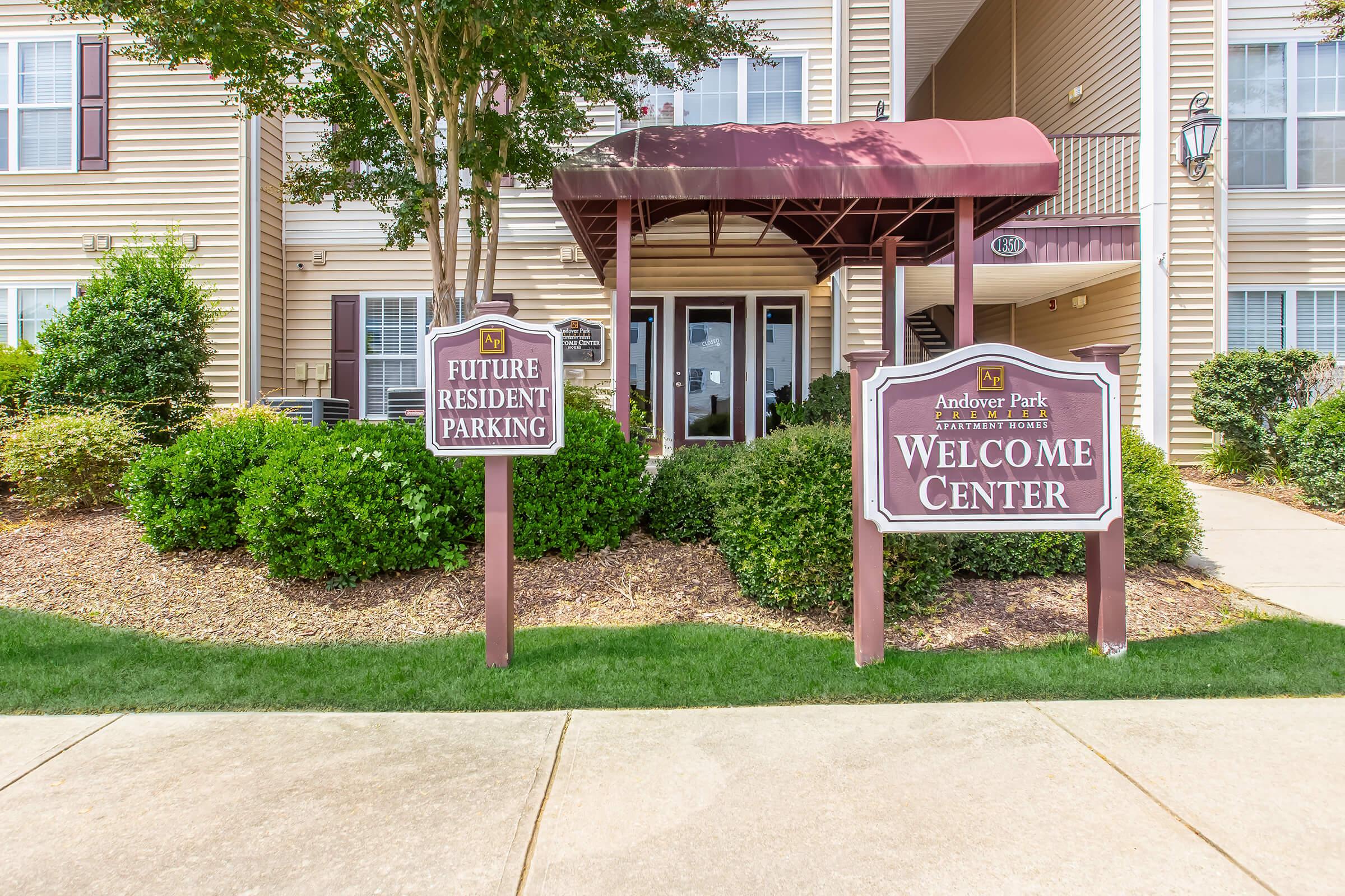 A welcoming entrance to Andover Park, featuring signs for "Future Resident Parking" and a "Welcome Center." The area is neatly landscaped with shrubs and small trees, creating an inviting atmosphere. The building in the background is light-colored with a maroon awning over the entrance.