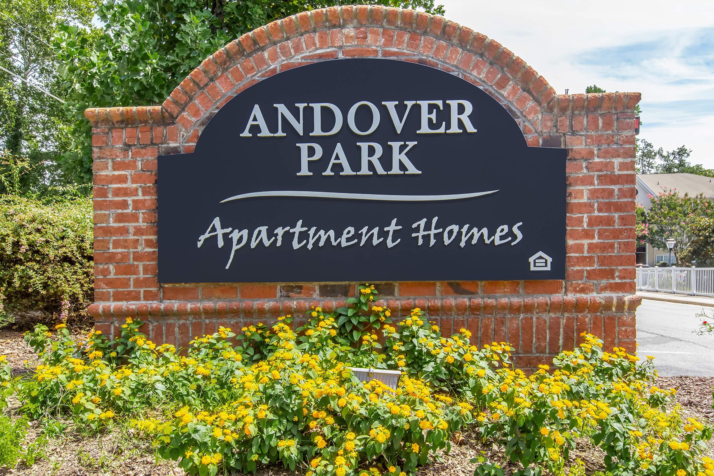 Sign for Andover Park Apartment Homes with a brick arch and colorful yellow flowers at the base. The sign features the name "Andover Park" prominently at the top and "Apartment Homes" below, set against a dark background. Lush greenery is surrounding the sign.