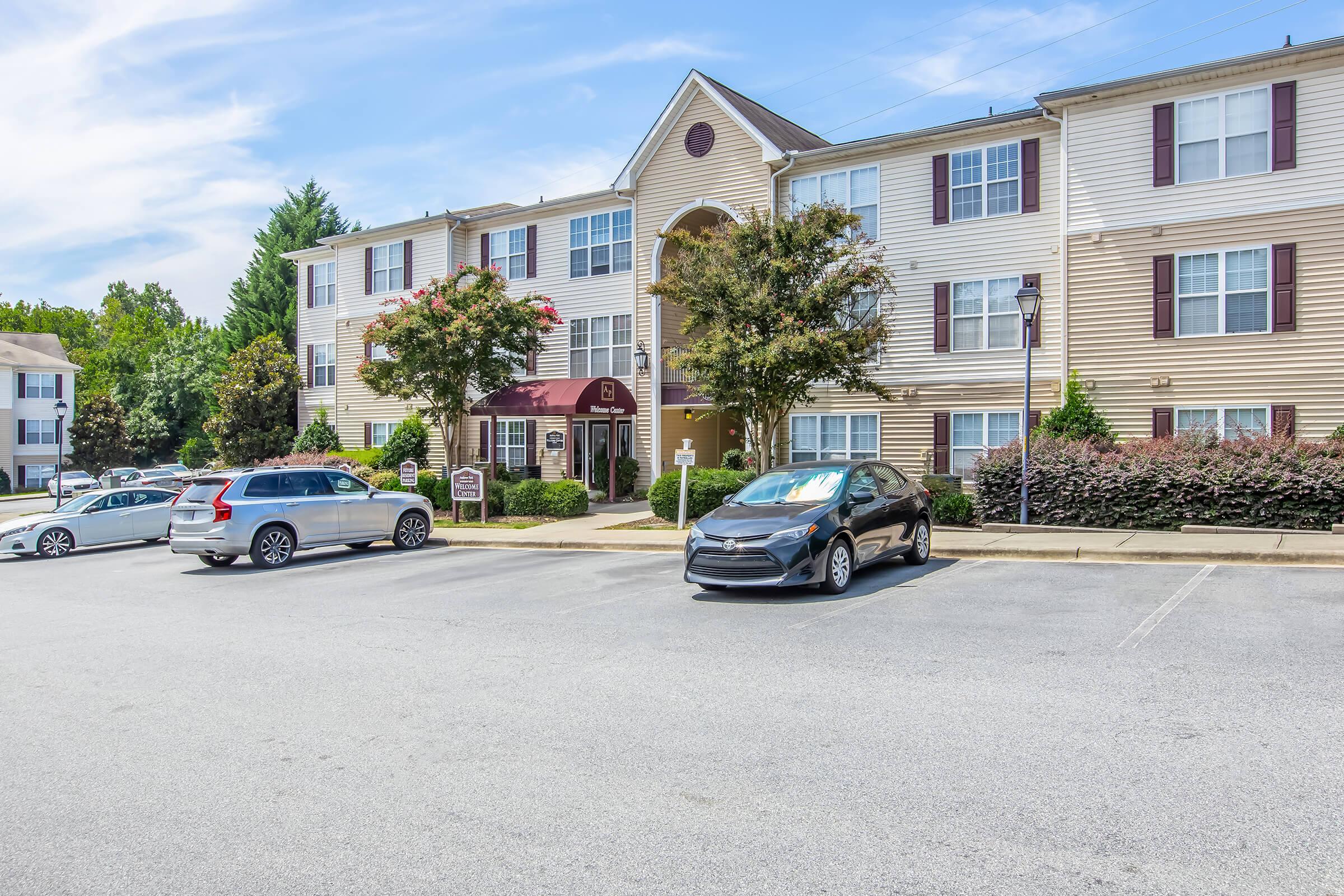 A three-story residential apartment building with a maroon awning at the entrance. Surrounding the building are landscaped areas with trees and shrubs. Several parked cars are visible in the foreground, with a light-colored SUV and a black sedan prominently displayed.