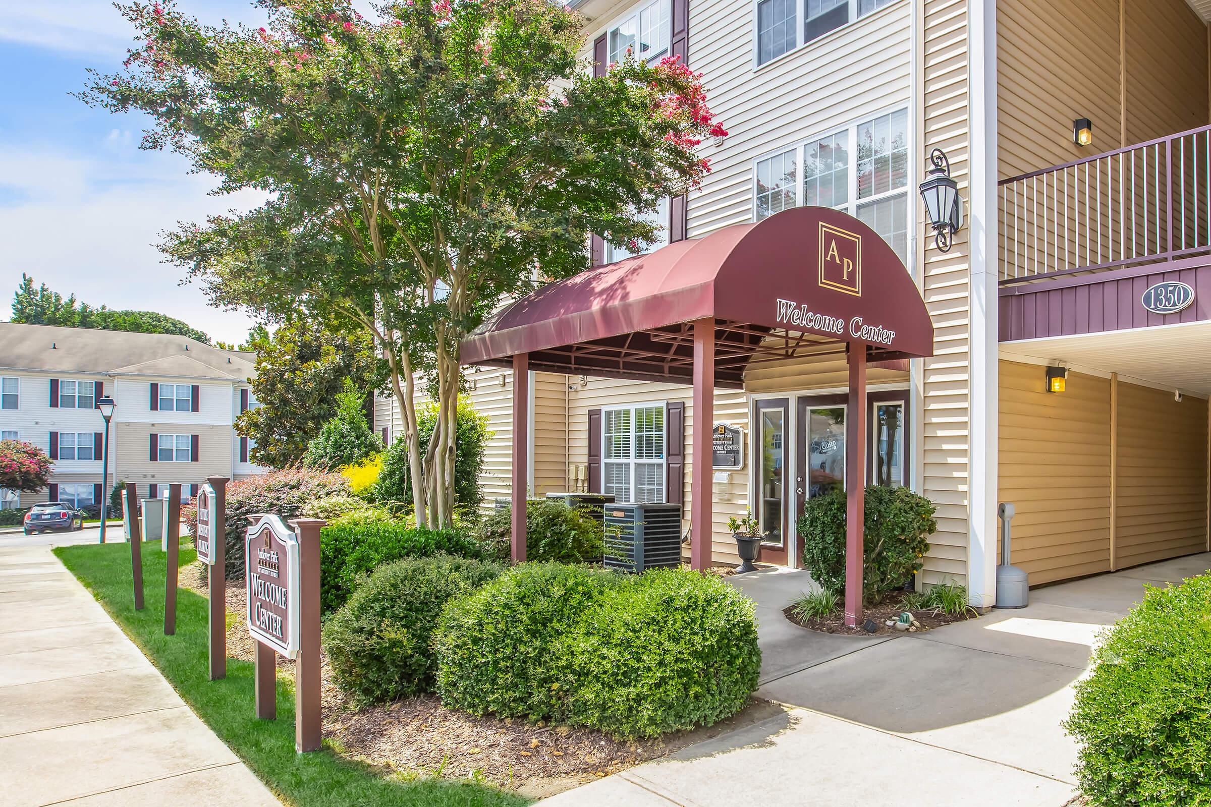 A welcoming entrance to an apartment complex featuring a covered canopy with a sign that reads "Welcome Center." The pathway is lined with manicured shrubs, and the building has multiple windows and a tan exterior. There's a clear blue sky with some clouds in the background.