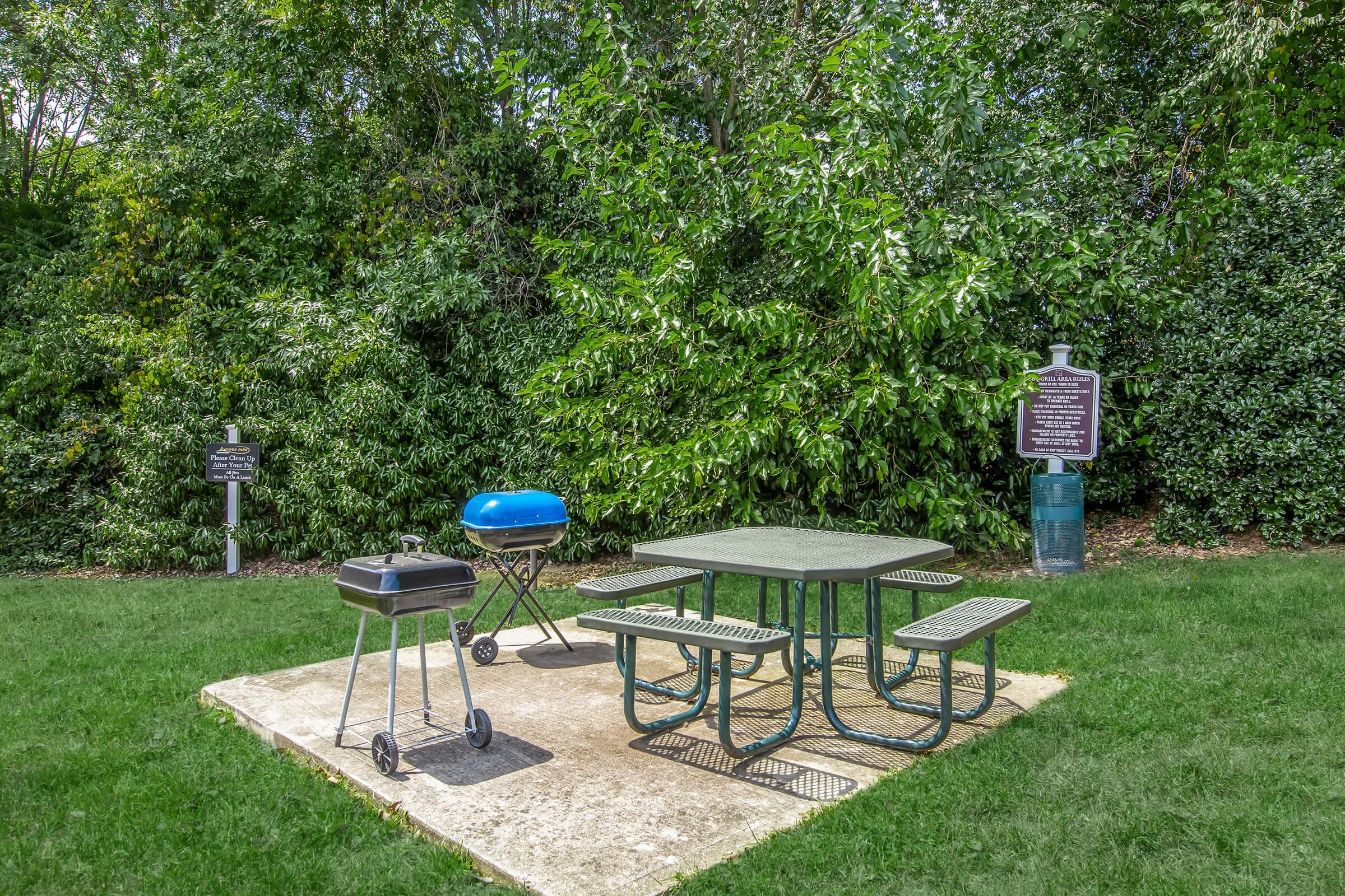 A green picnic table surrounded by four benches on a concrete slab, with a small charcoal grill nearby. The background features lush greenery and trees. A sign can be seen on the left, indicating information about the area. The scene is set in a park-like environment.