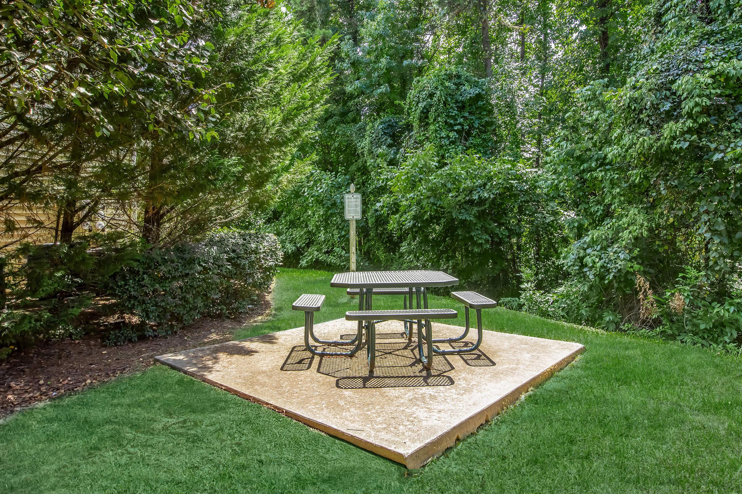 A black metal picnic table with attached benches sits on a concrete pad surrounded by lush green foliage and grass in a secluded outdoor area. The setting is tranquil, with trees providing shade and a signpost visible in the background.
