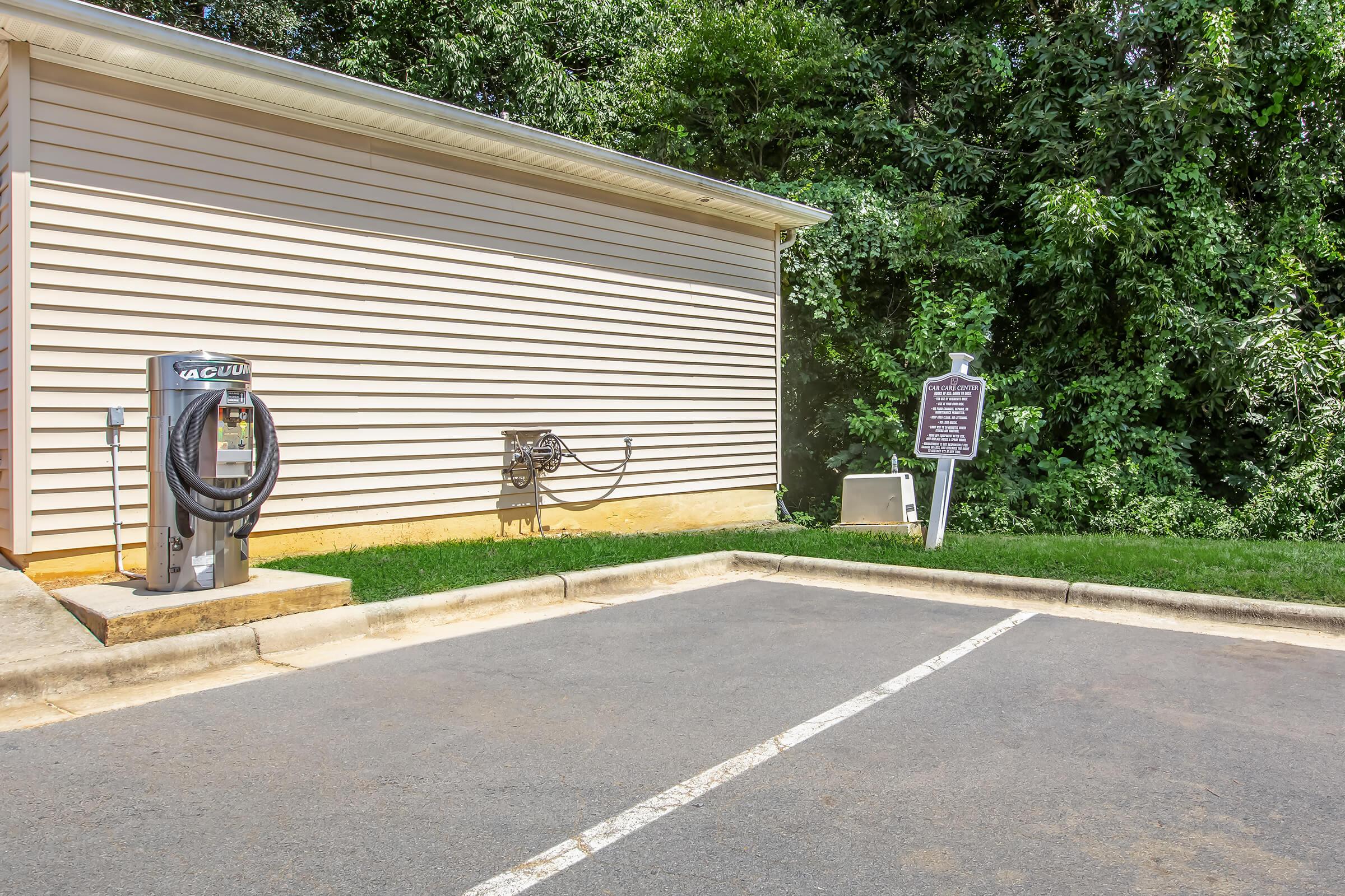 A vacuum station with hoses stands beside a building, located in a parking area surrounded by green vegetation. A sign is positioned nearby, and the pavement is marked with a single parking line.