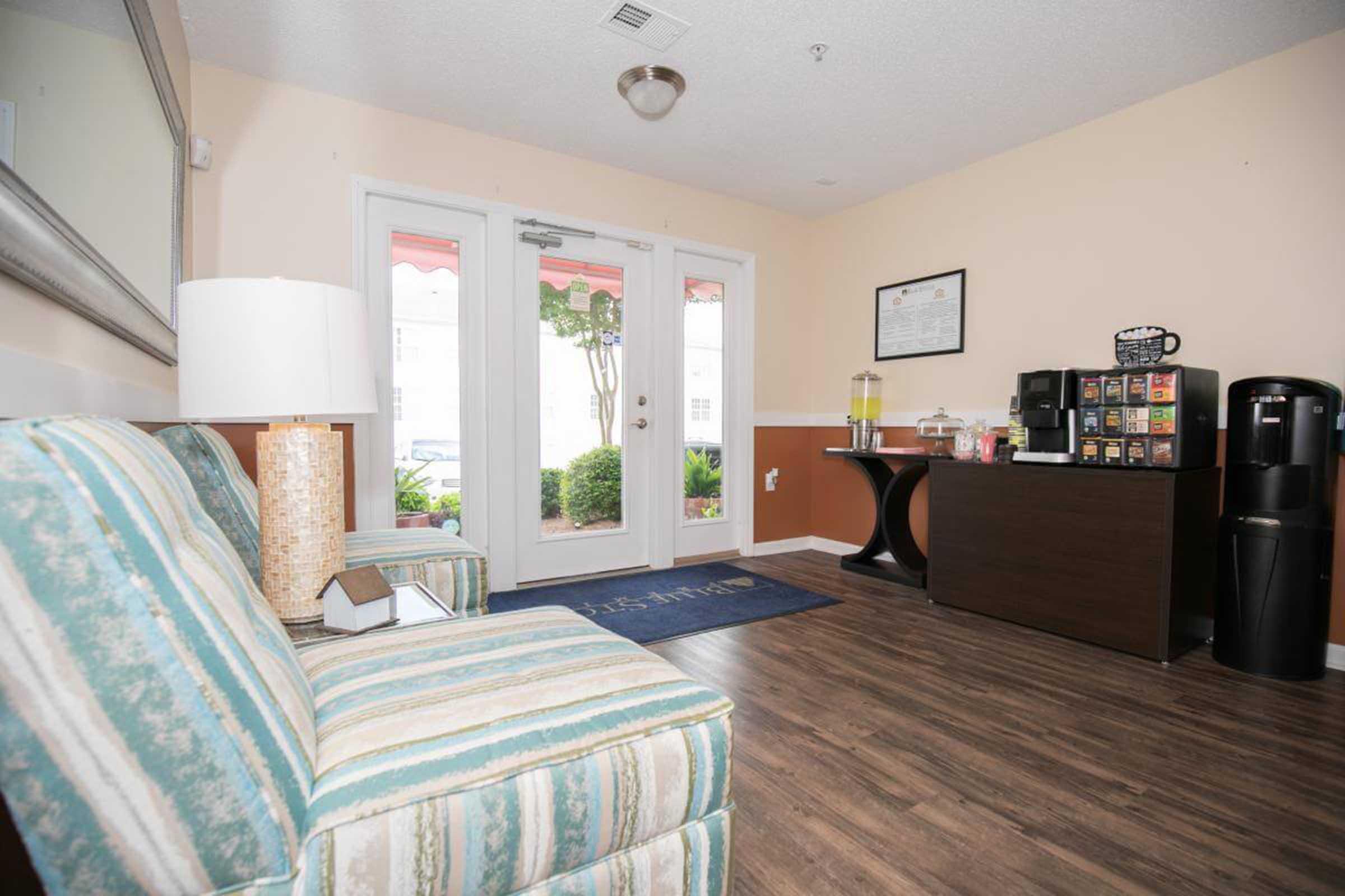 A cozy lobby area featuring a striped sofa, a table with refreshments, a coffee machine, and a water dispenser. There are glass doors leading outside, framed by plants, and a decorative lamp on a side table. The floor is wooden, and the overall ambiance is welcoming and comfortable.