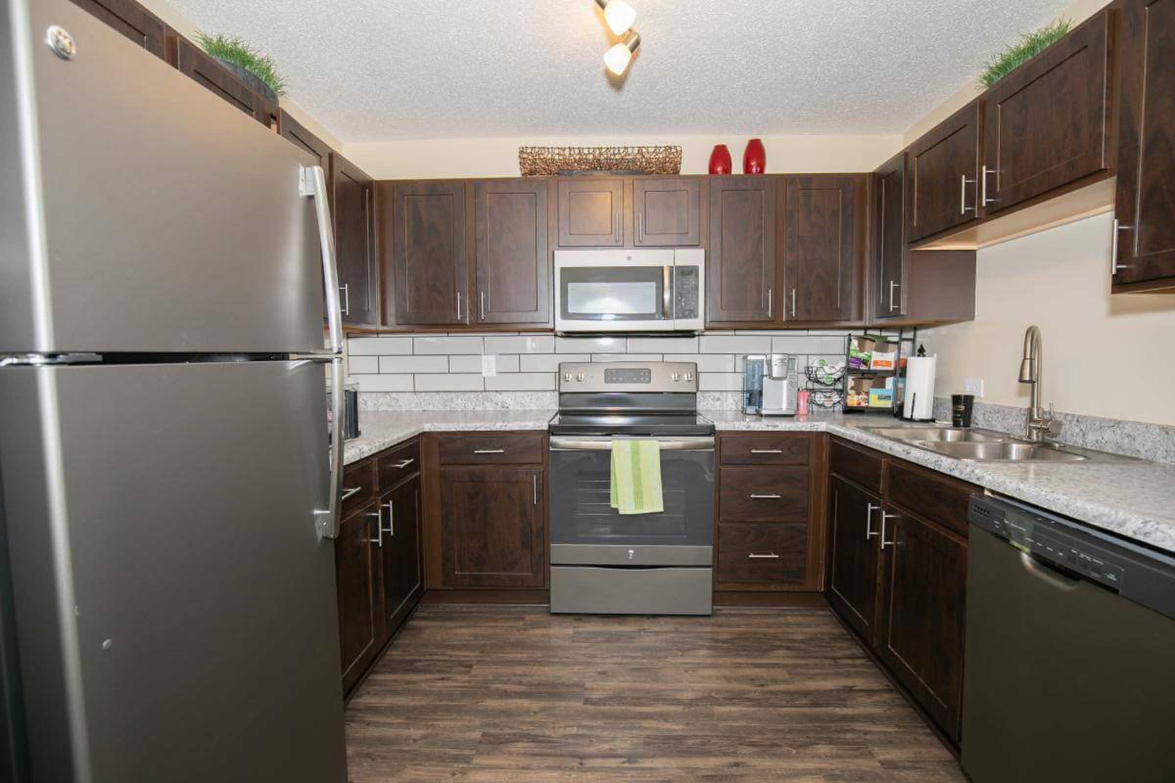 Spacious modern kitchen featuring dark wood cabinets, stainless steel appliances, and a granite countertop. Includes a refrigerator, oven, microwave, and sink. The decor is complemented by plants and red accents, with a clean and contemporary design. Perfect for cooking and entertaining.