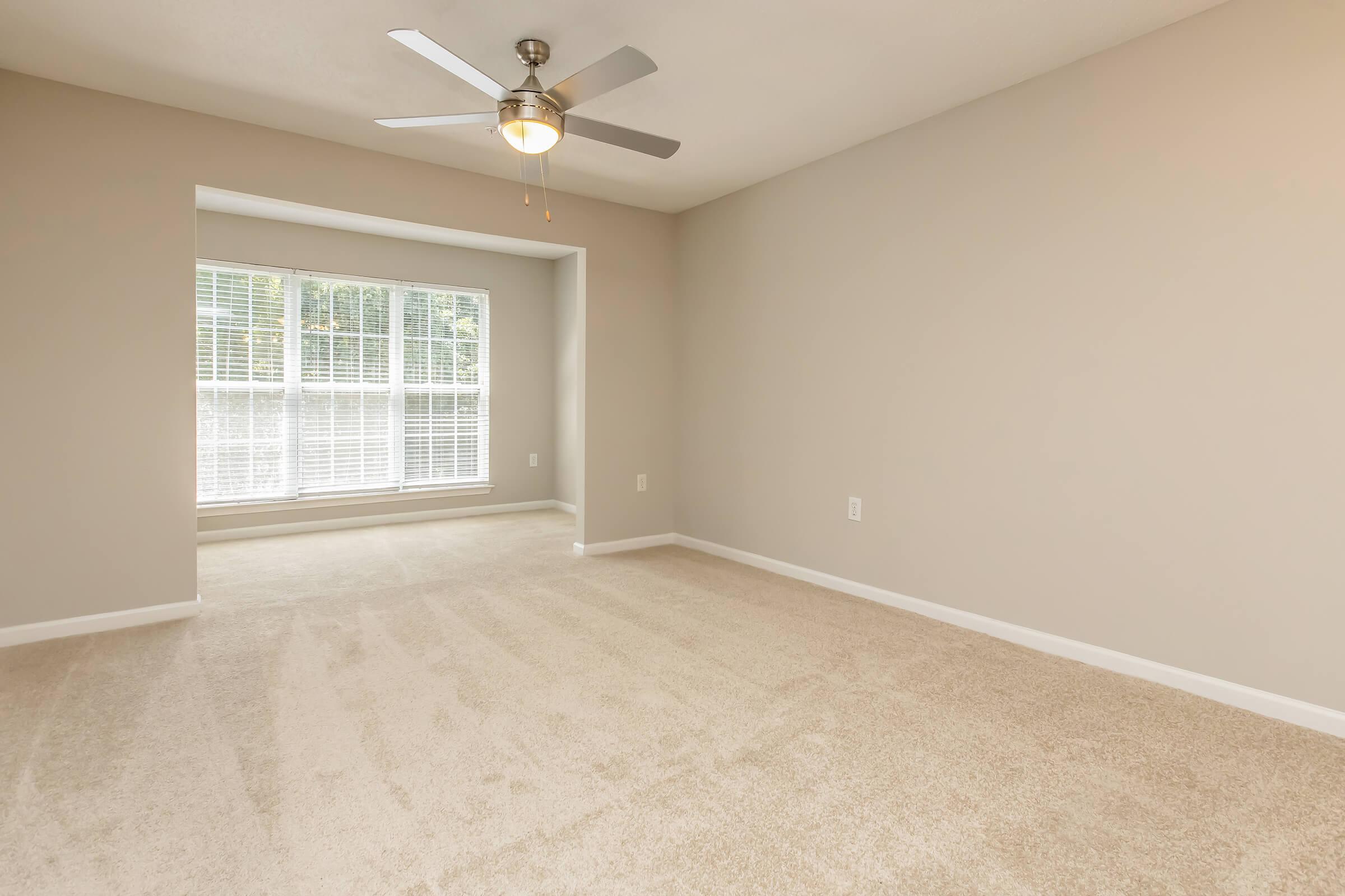 A spacious room with light beige walls and carpet, featuring a ceiling fan. A large window with blinds allows natural light to fill the space, showcasing a view of the outdoors. The area is uncluttered and ready for furniture arrangement.