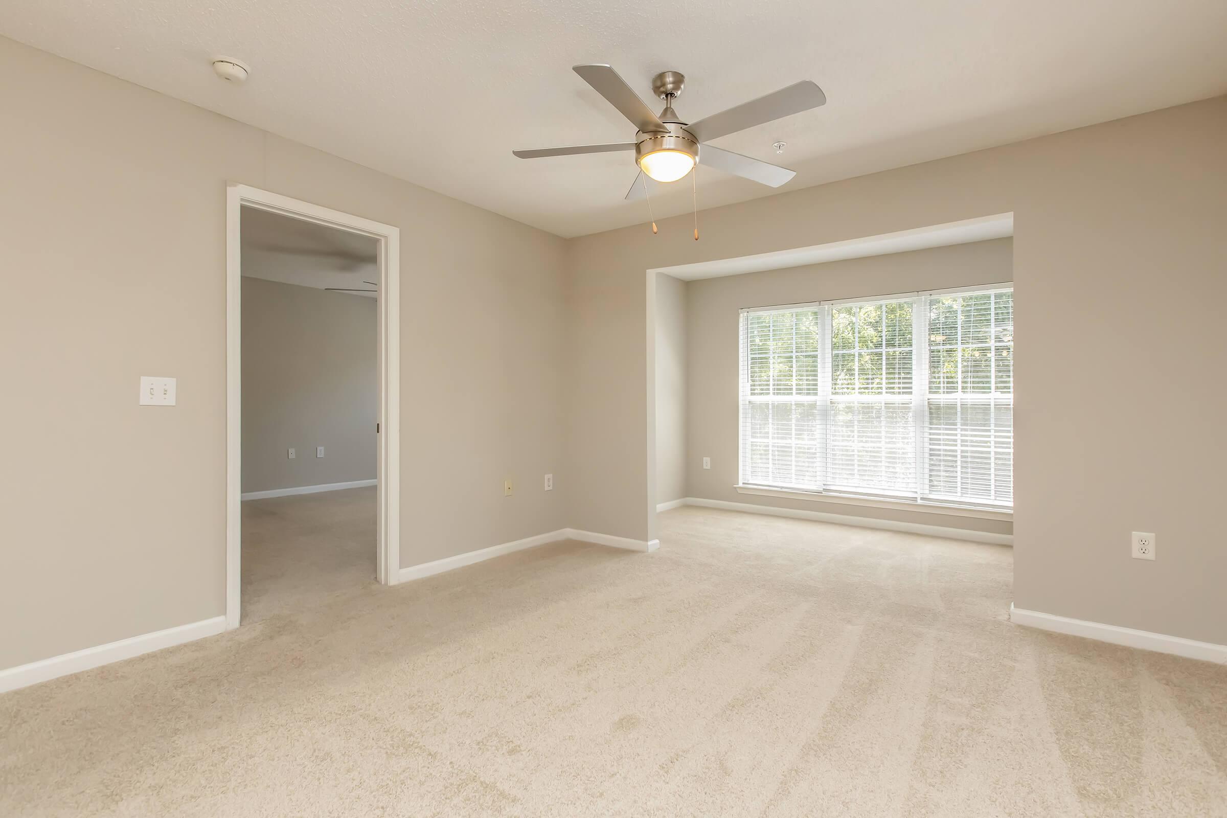 A spacious and well-lit living room featuring light beige walls and plush carpet. A ceiling fan is mounted above, and large windows allow natural light to fill the room. The room includes an open doorway leading to another area, enhancing its airy feel.