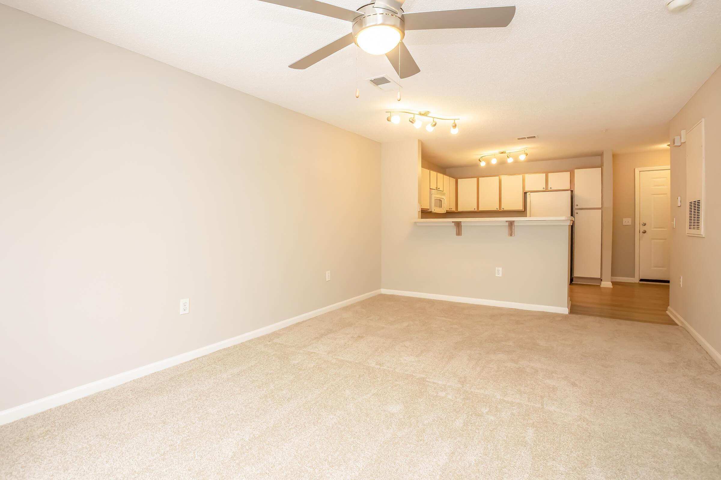 A spacious, undecorated living area featuring light beige walls and soft carpet. A ceiling fan is mounted above, and the kitchen is visible in the background with white cabinetry, appliances, and a countertop. The open layout creates a welcoming and bright atmosphere.