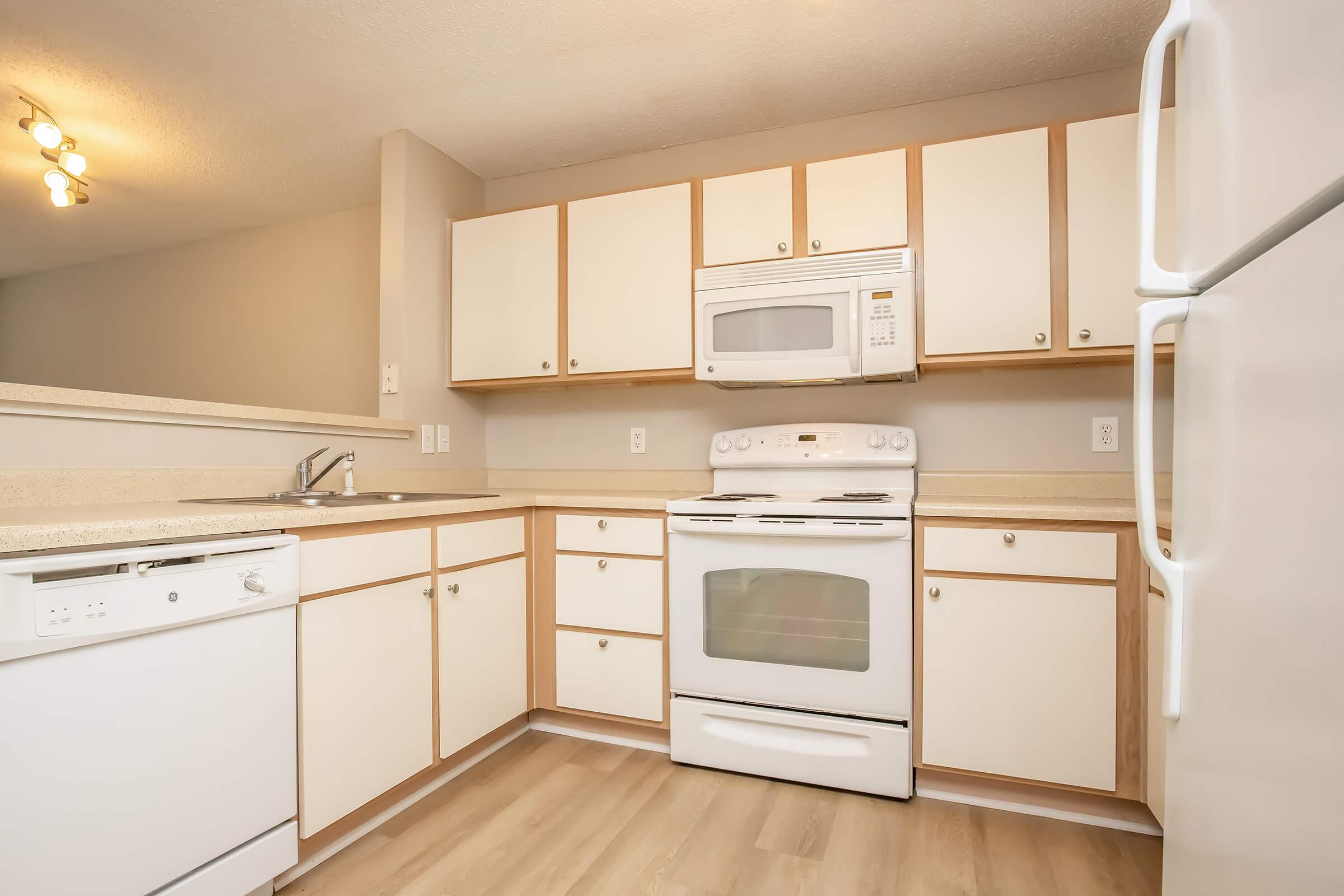 Bright, modern kitchen featuring white cabinetry with light wood accents, a double sink, a refrigerator, microwave, and an oven/stove combination. The countertop is light-colored, and the flooring appears to be laminate. Soft lighting from overhead fixtures creates a warm ambiance.