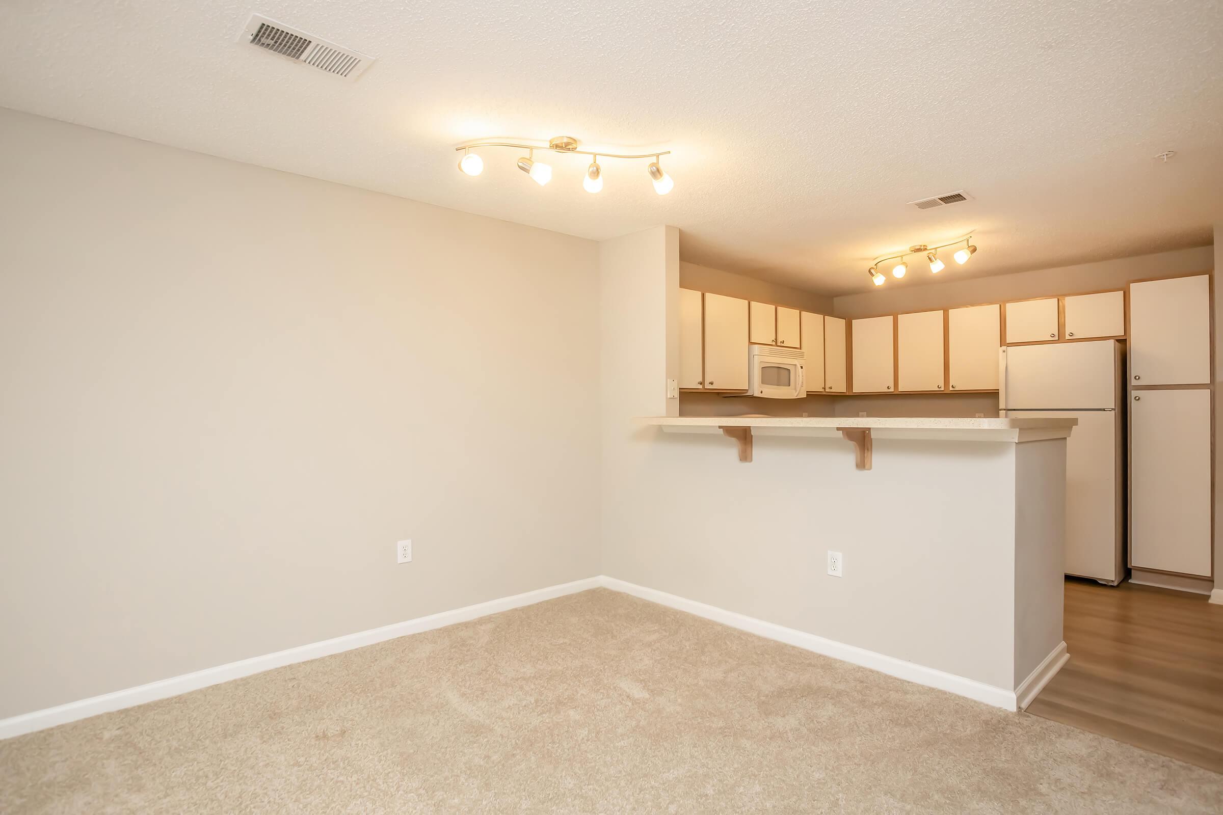A spacious room with light beige walls and carpet flooring. On one side, there is a kitchen area with white cabinets, a microwave, and a refrigerator. The ceiling features modern light fixtures. The overall atmosphere is bright and inviting, suitable for a living area or dining space.