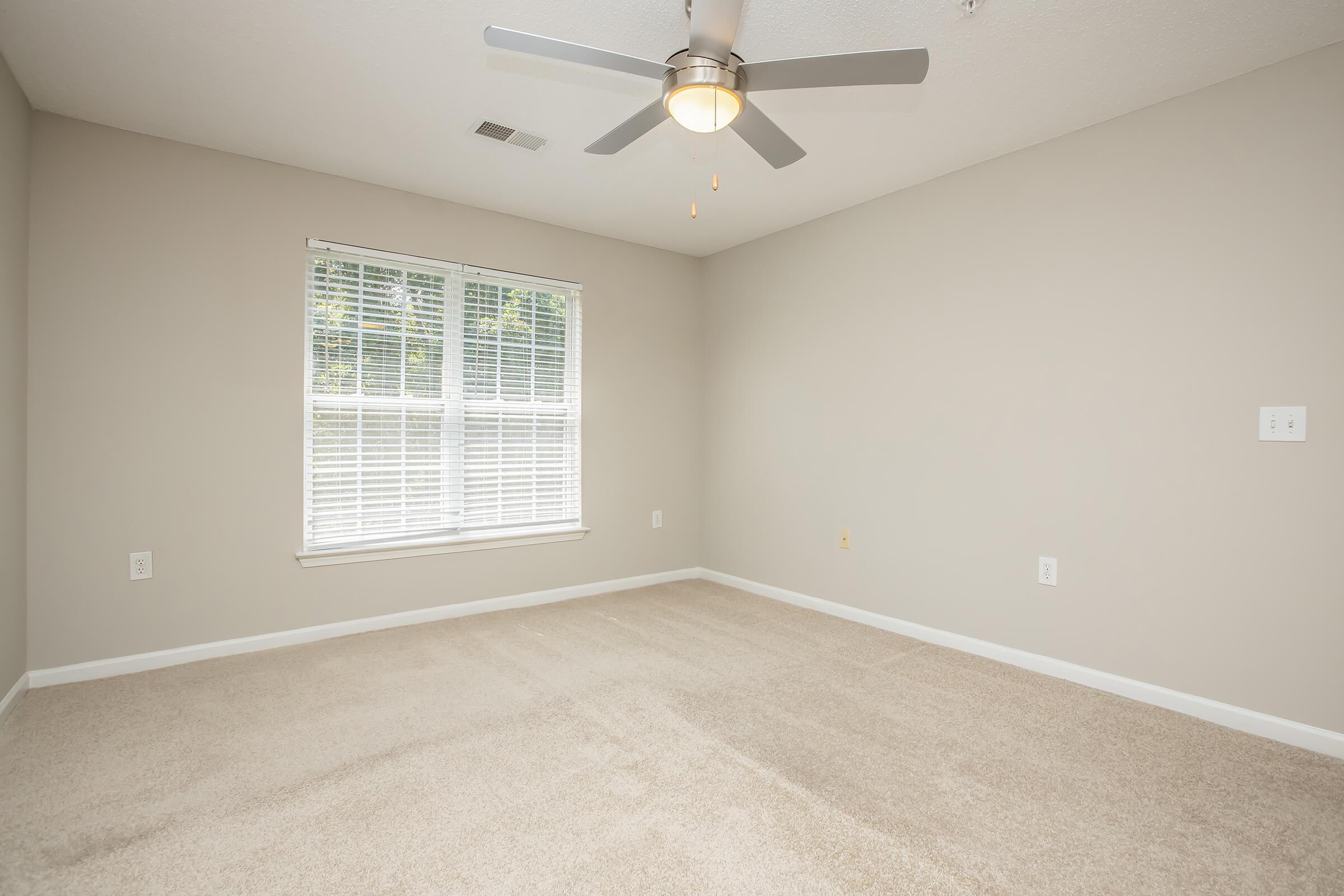 Empty room with light beige walls and a ceiling fan. The floor is covered in light-colored carpet. A window with white blinds allows natural light to enter, highlighting the spacious, uncluttered area.