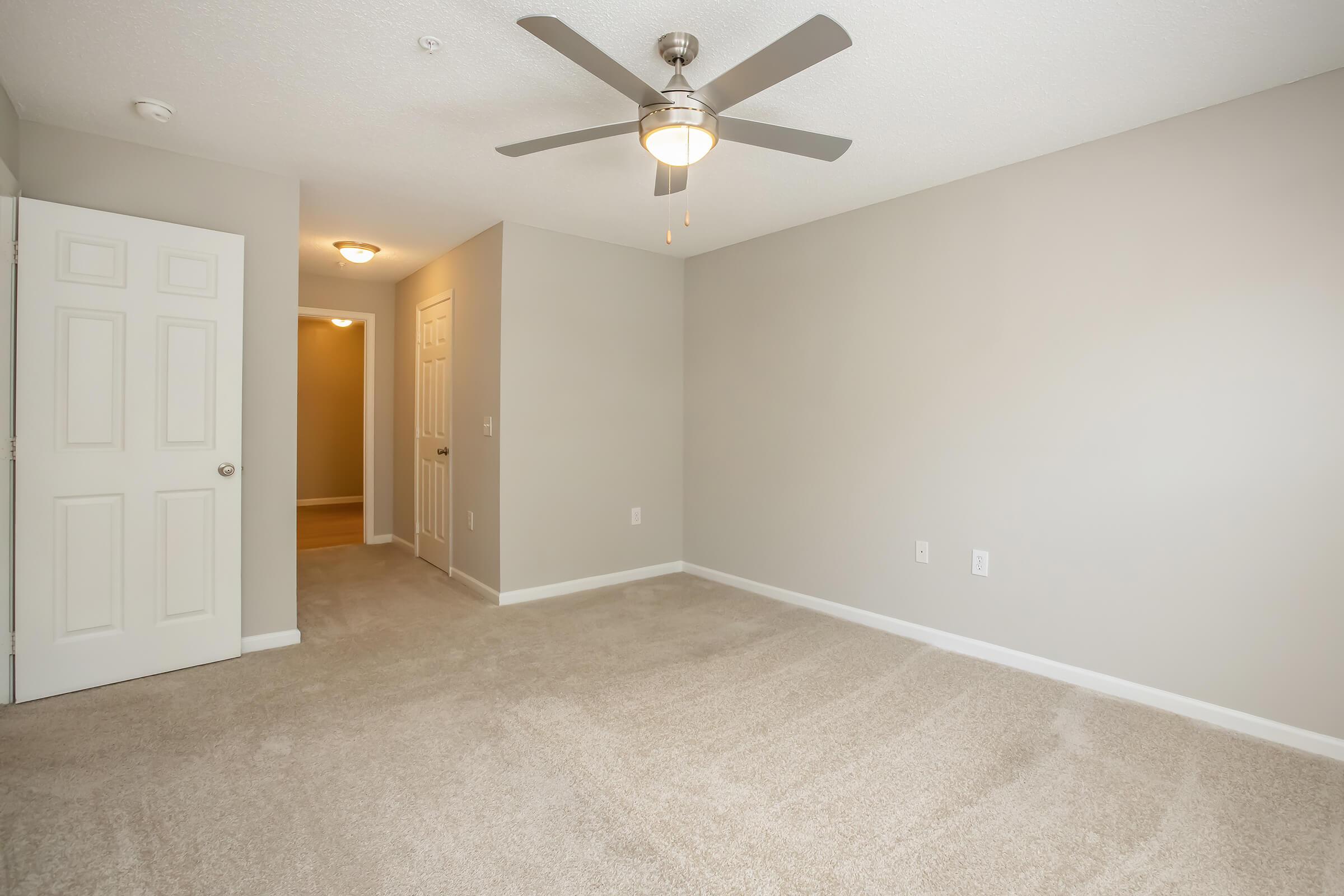 A spacious, empty room with light gray walls and beige carpet. It features a ceiling fan with a light, and a doorway leading to a hallway with additional doors. Natural light from the hallway brightens the space, creating a warm and inviting atmosphere.