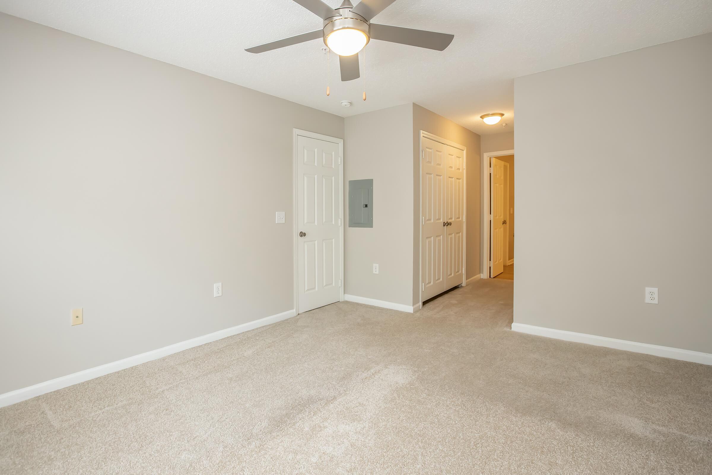 Spacious, neutral-colored room with plush, light beige carpeting. A ceiling fan is mounted above, and there's a door leading to a hallway with additional doors. The walls are painted a soft gray, creating a calm atmosphere. The room is well-lit, highlighting its open layout.
