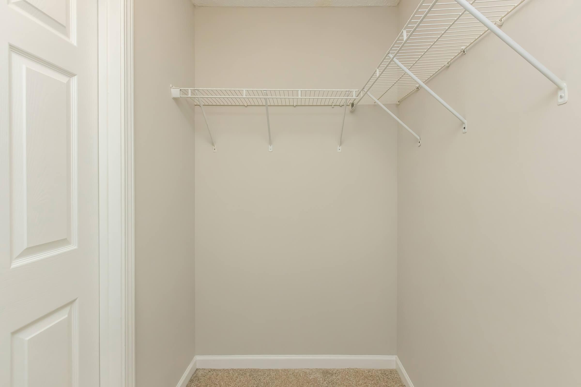 Empty closet featuring light beige walls and carpeted flooring. A white wire shelf is mounted on one wall with several brackets, providing storage space, while the opposite wall has a closed white door. The overall space is clean and uncluttered, suitable for organization.