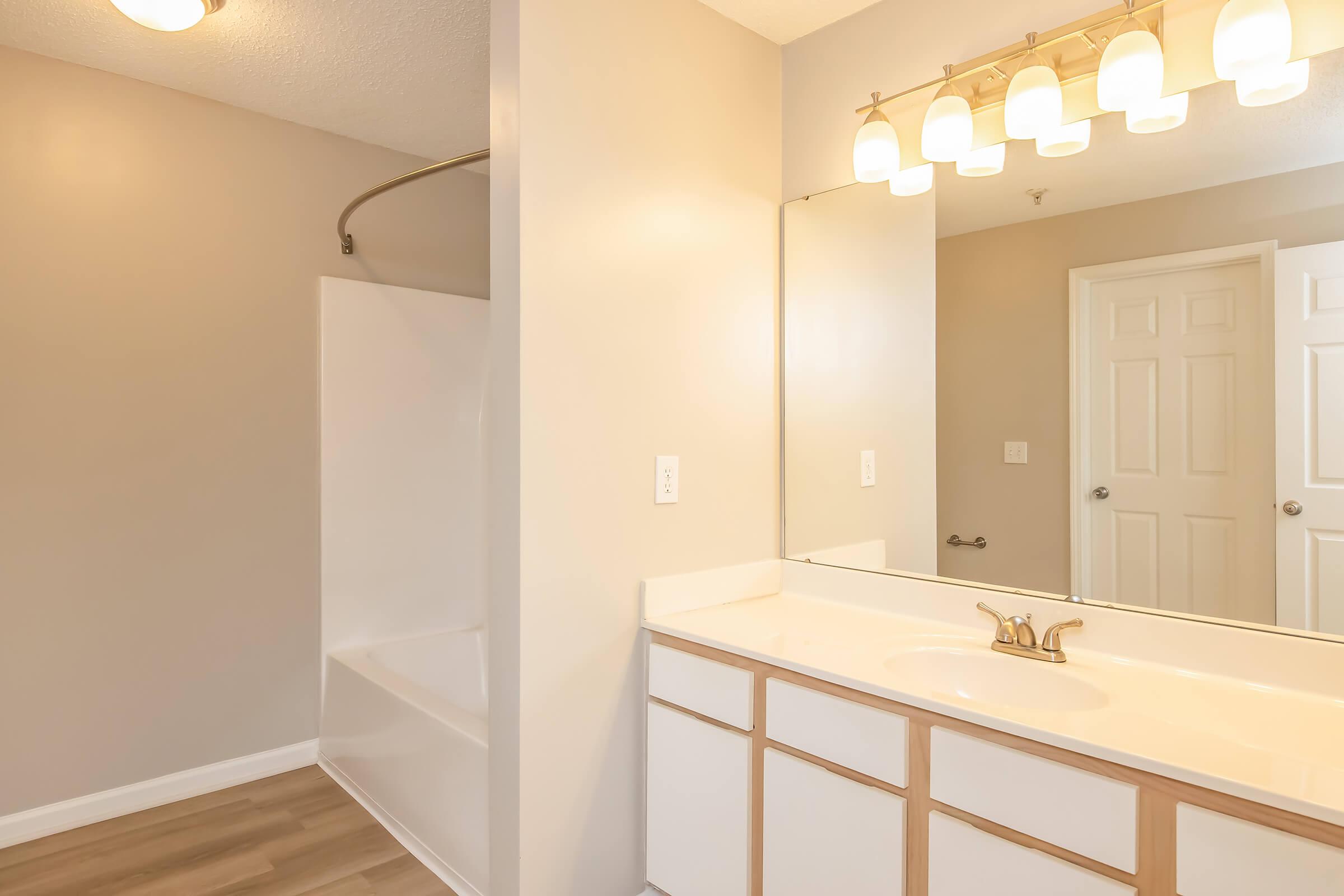 A well-lit bathroom featuring a shower with a curved curtain rod, a single sink vanity with a light fixture above, neutral wall colors, and a door leading to another room. The floor has a light wood finish, and the space appears clean and modern.