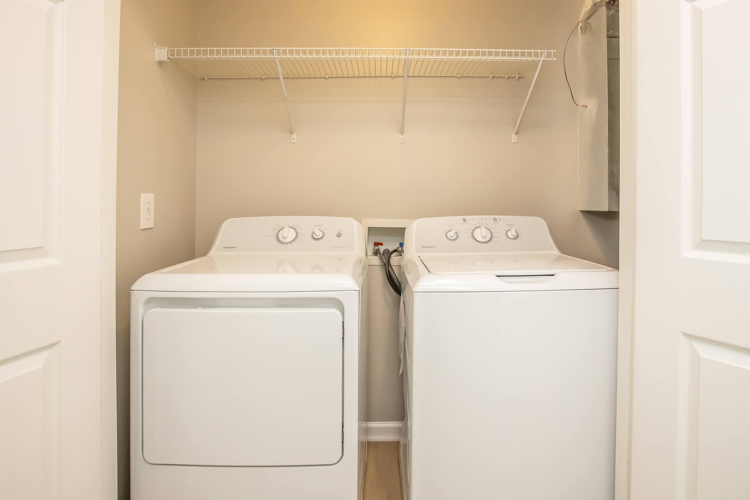 A bright laundry room featuring a stacked washer and dryer side by side in a neutral-colored space. Above the appliances, there's a wire shelf for storage. The walls are painted a light color, and there are two white doors leading into the room.