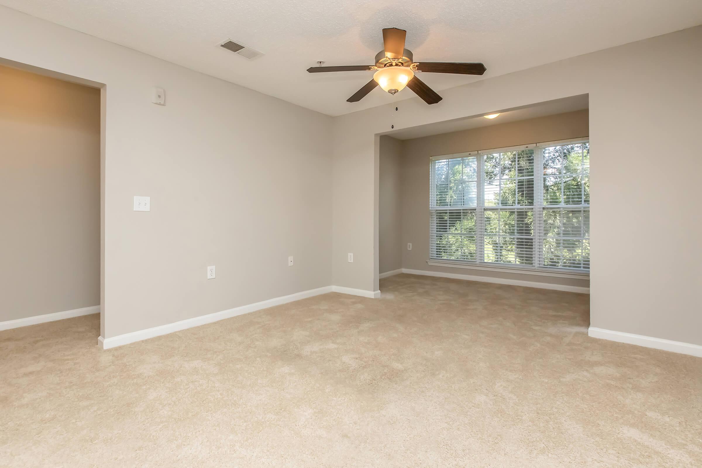 A spacious, empty room with beige carpet and light gray walls. It features a ceiling fan with a light fixture, a large window allowing natural light, and two openings leading to separate areas. The overall ambiance is bright and airy, suitable for various uses.