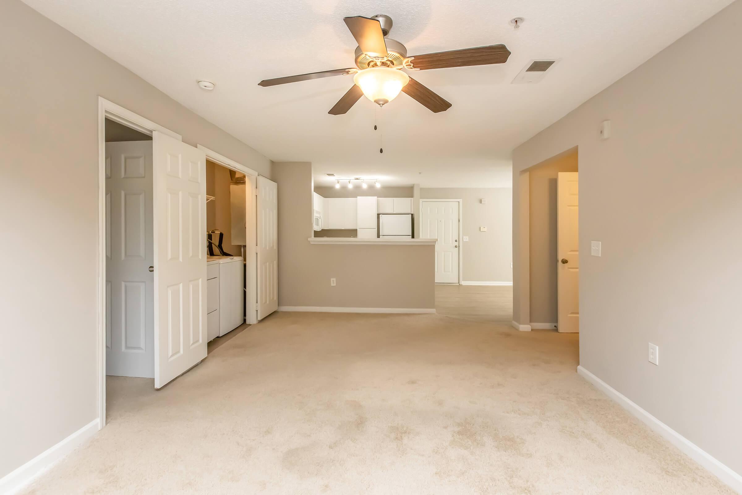 A spacious living area with beige carpet flooring and light gray walls. A ceiling fan hangs above, adding a cozy touch. In the background, a kitchen area is visible with an open layout, and laundry facilities can be seen through the partially open double doors. Soft natural light fills the space.