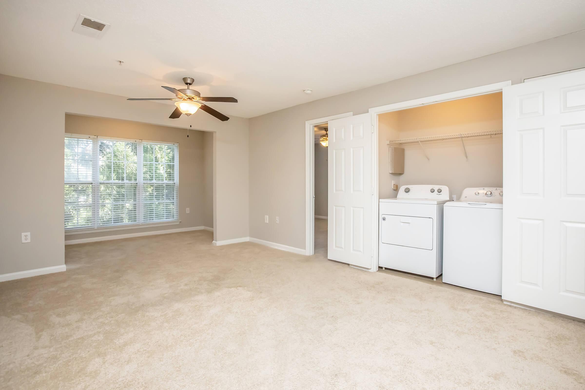 Bright and spacious room with light beige carpet, featuring a ceiling fan. Large windows provide natural light. To one side, there is a closet area with a washer and dryer, with double doors for access. The walls are painted in a soft neutral color, creating a warm and inviting atmosphere.