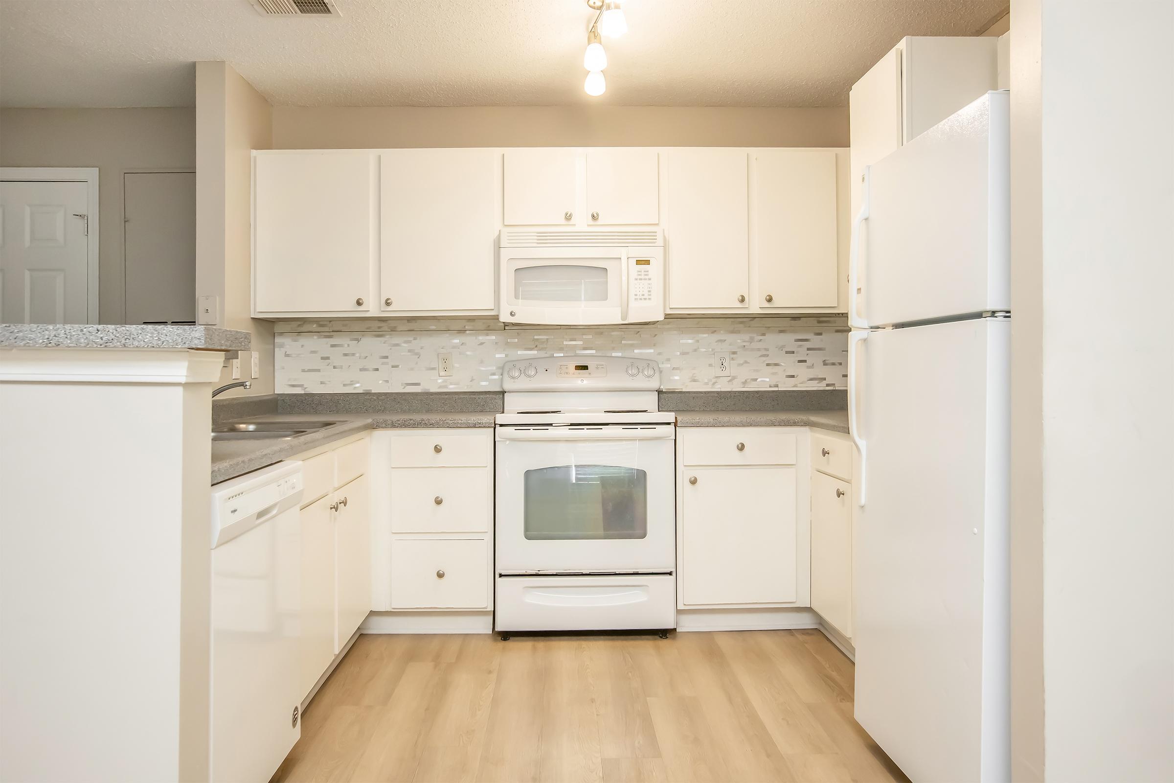 Modern kitchen featuring white cabinetry, a countertop, and stainless steel appliances. Includes a dishwasher, microwave, stove, and refrigerator. The backsplash consists of mosaic tiles, and the flooring is a light wood finish. Bright lighting fixtures hang from the ceiling, enhancing the spacious feel.