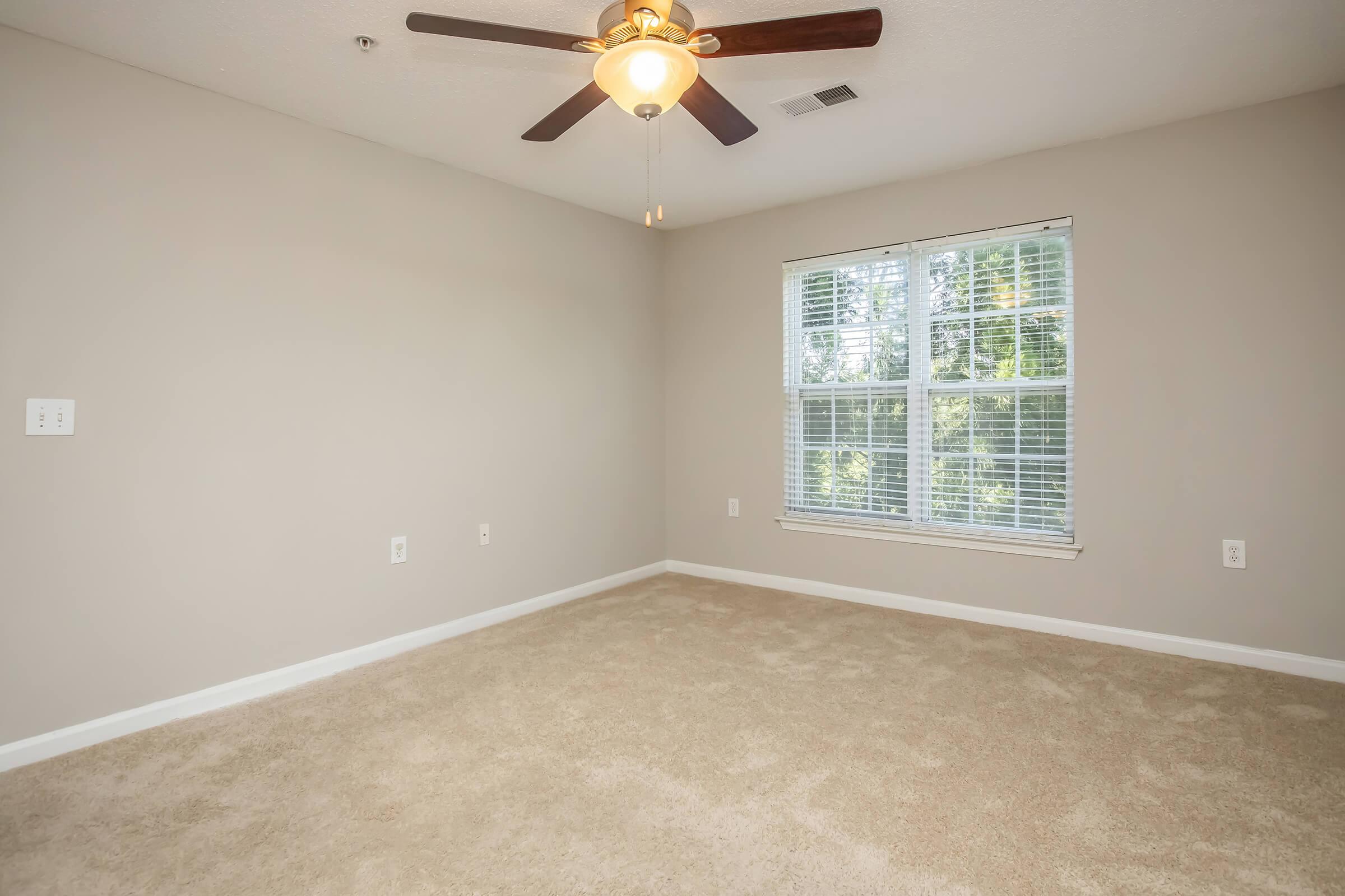 A spacious, empty room featuring light beige carpet, a ceiling fan with wood blades, and a large window with white blinds allowing natural light to enter. The walls are painted a soft, neutral color and the room has a clean, uncluttered appearance.
