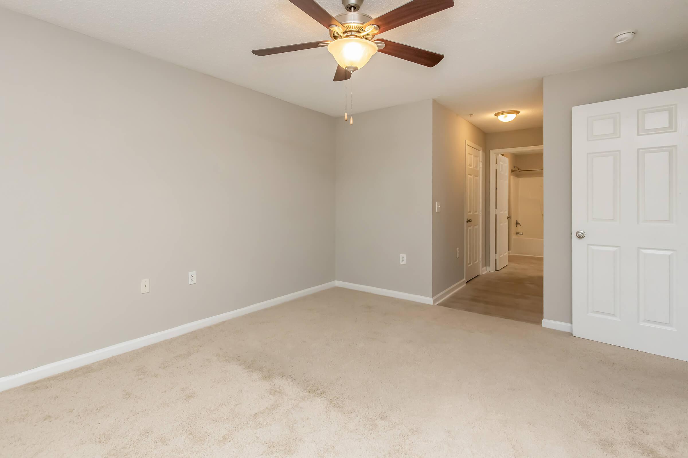 An empty room featuring light gray walls, carpeted flooring, a ceiling fan with a light fixture, a door leading to a bathroom, and another door to a closet. The space is well-lit, appearing clean and spacious, with no furniture present.