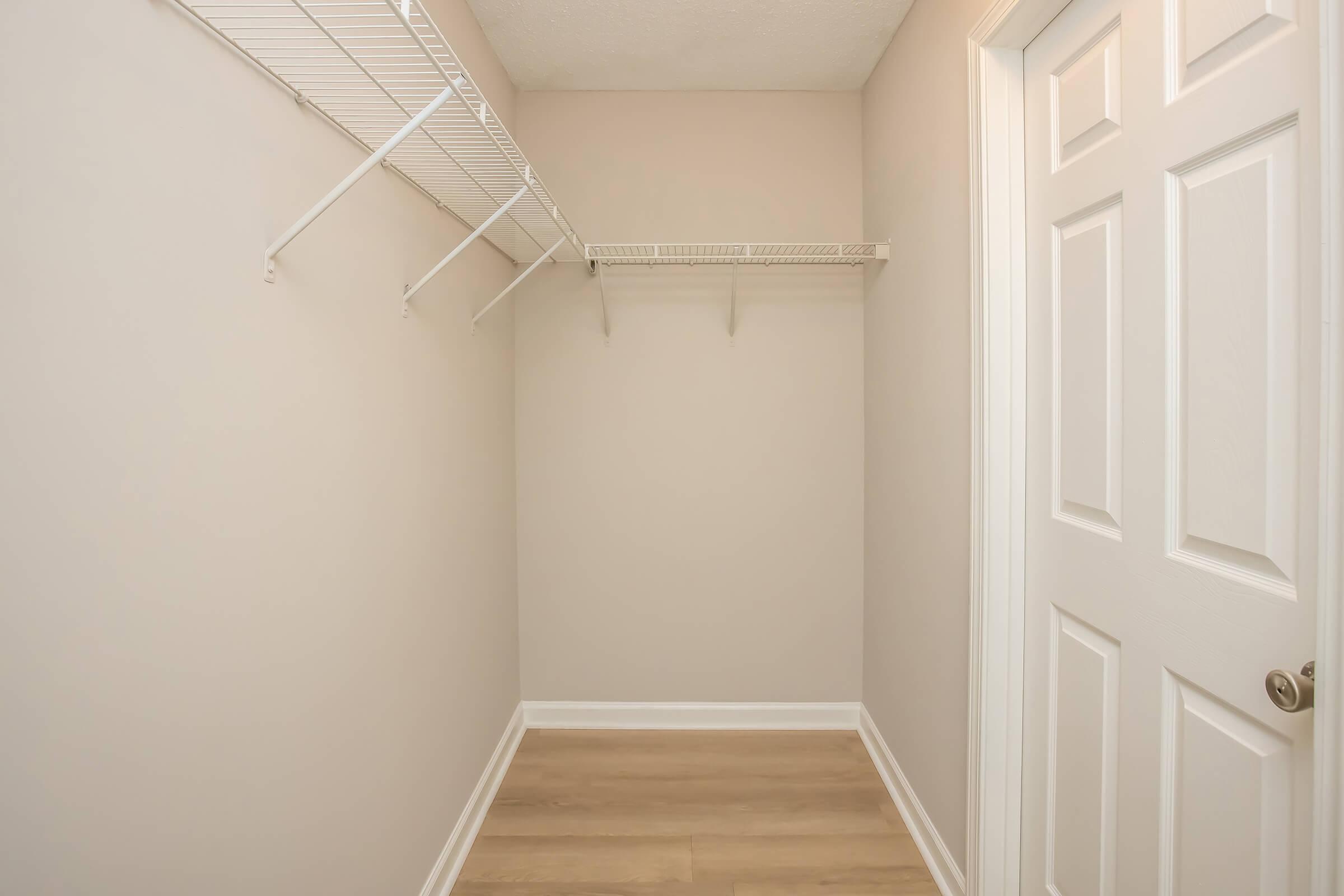 Empty closet with beige walls and a light wooden floor. A white wire shelving unit is attached to one wall, and a closed white door is visible on the right side. The space is well-lit and minimalistic, providing a clear view of the uncluttered interior.