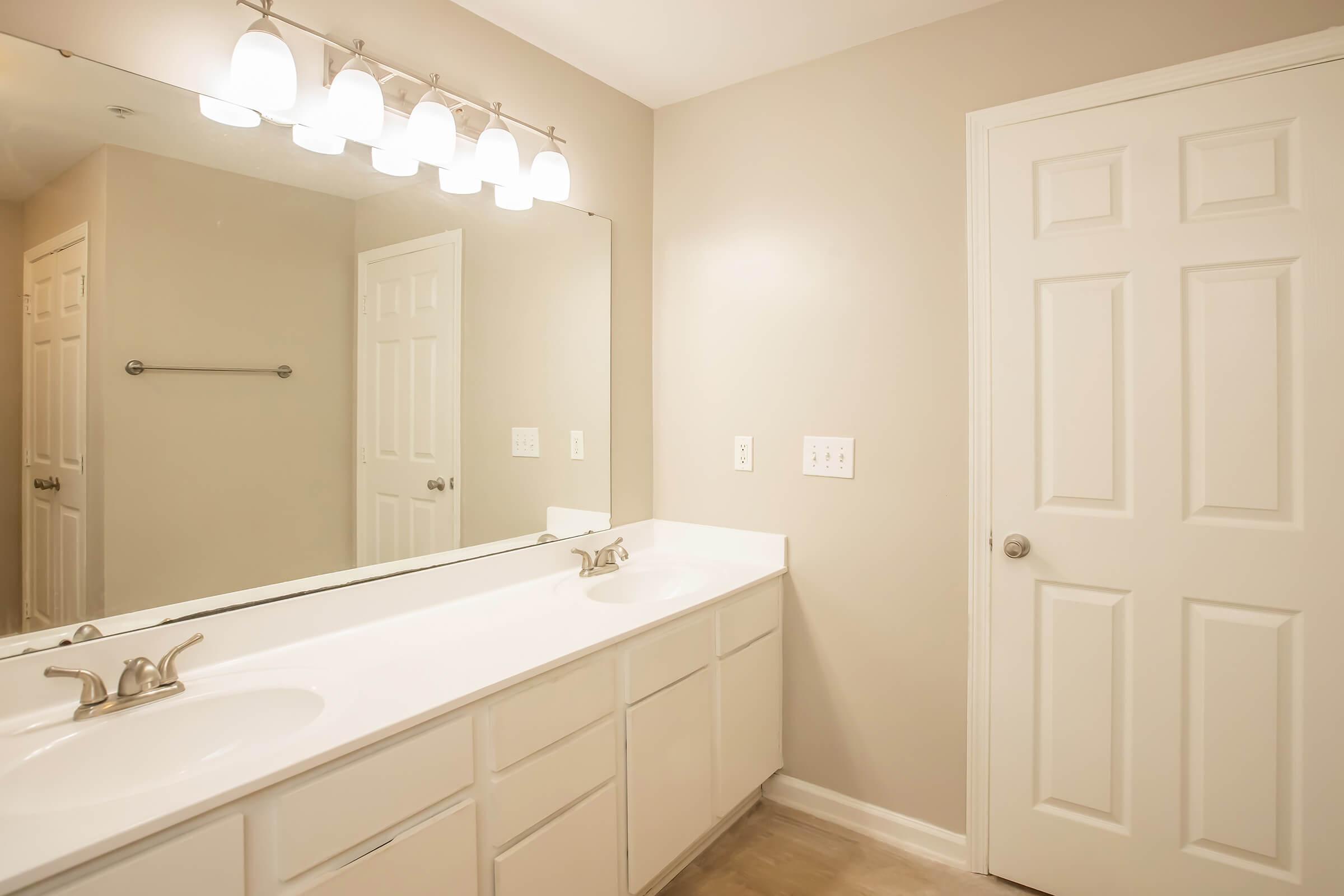 Bright and modern bathroom featuring a double vanity with a white countertop, two sinks, and white cabinetry. A large mirror spans the length of the vanity, reflecting the room's light. The walls are painted a soft beige, and there's a closed white door and towel rack on the adjacent wall.