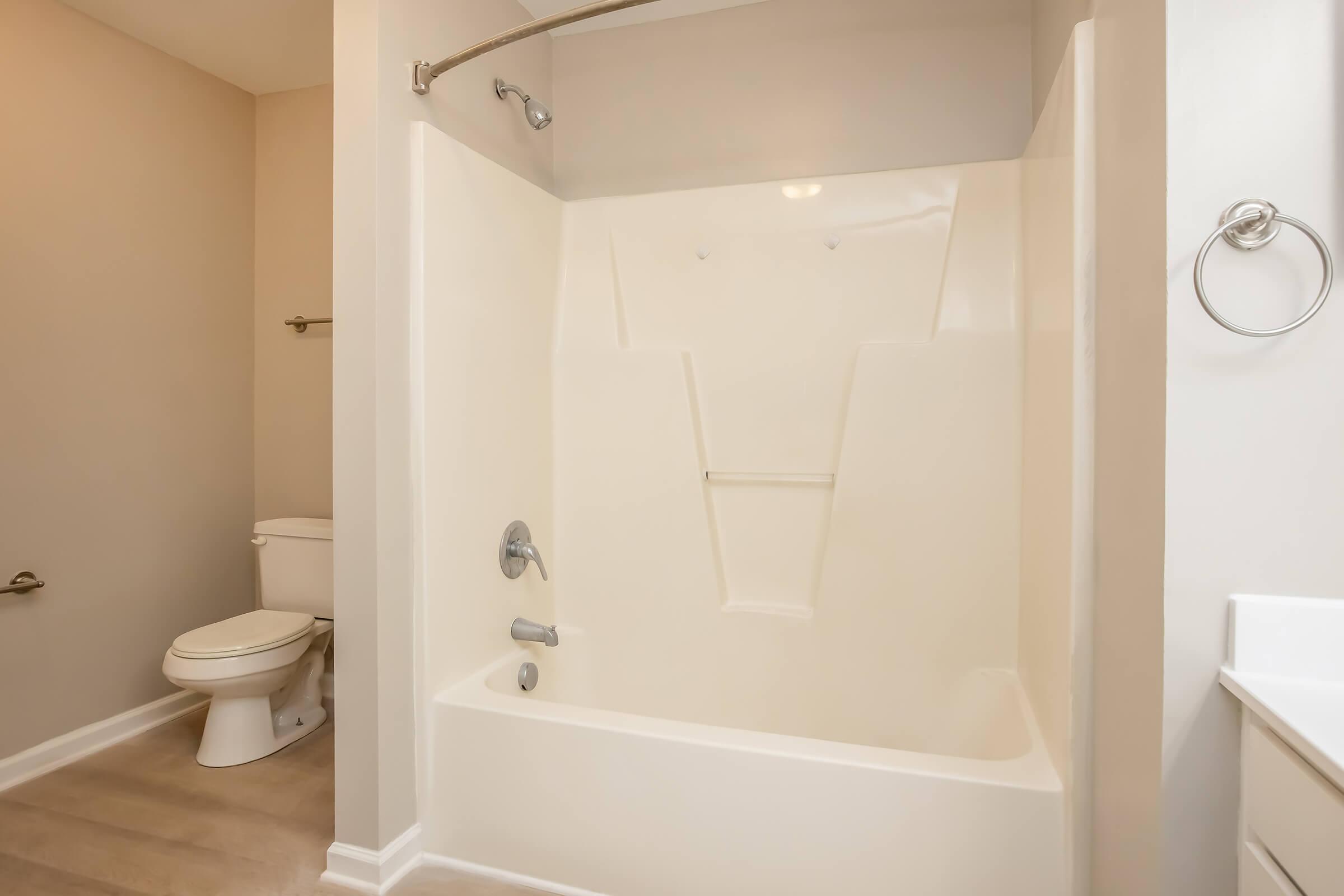 A clean and modern bathroom featuring a white bathtub and shower combination. The walls are light-colored and there is a toilet in the background. A towel ring is mounted on the wall, and the flooring is light-colored laminate. Natural light brightens the space.