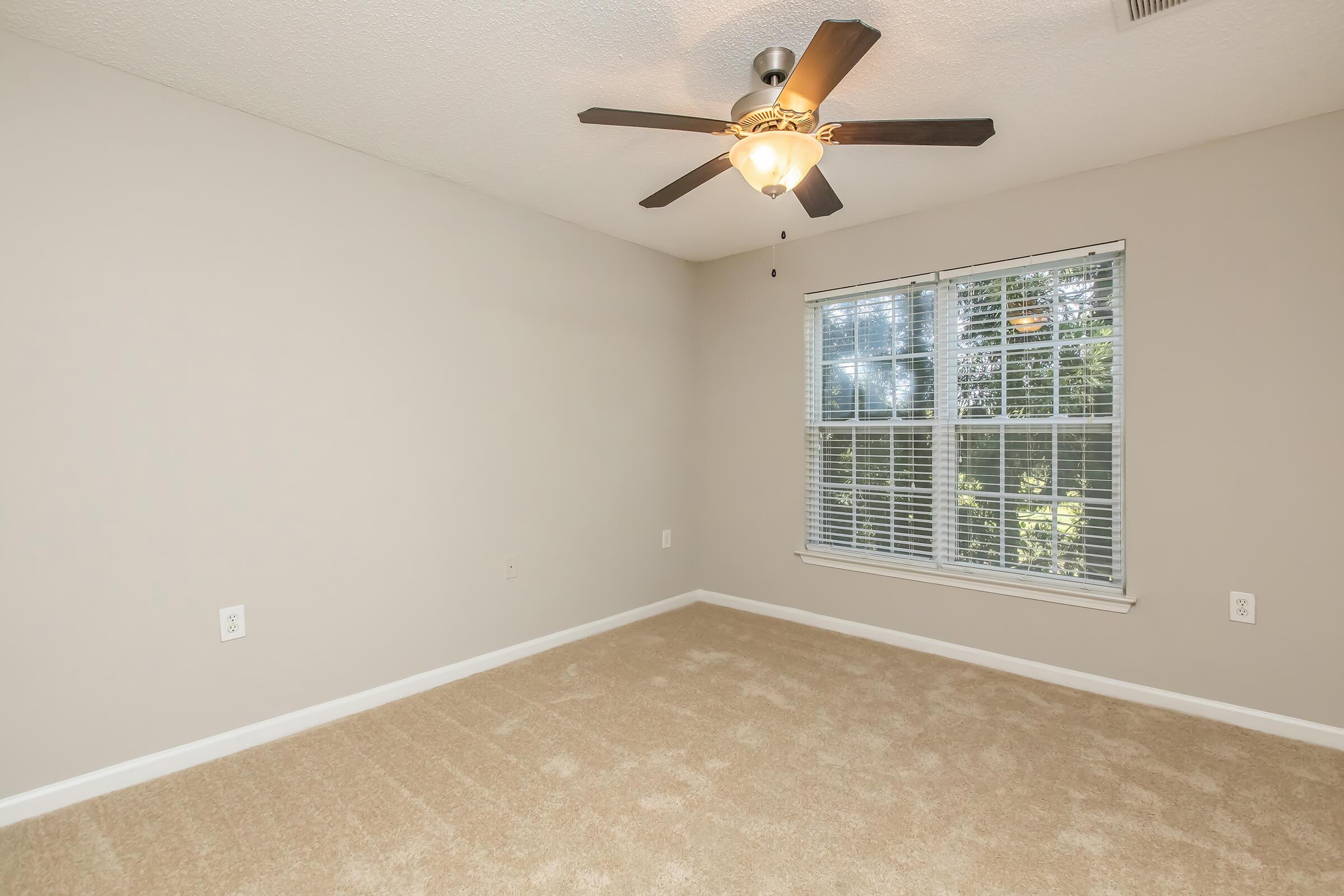 A vacant room with beige walls and light-colored carpet. It features a ceiling fan with a light fixture and a large window allowing natural light in, with blinds partially open. The space is empty, creating a bright and airy atmosphere.