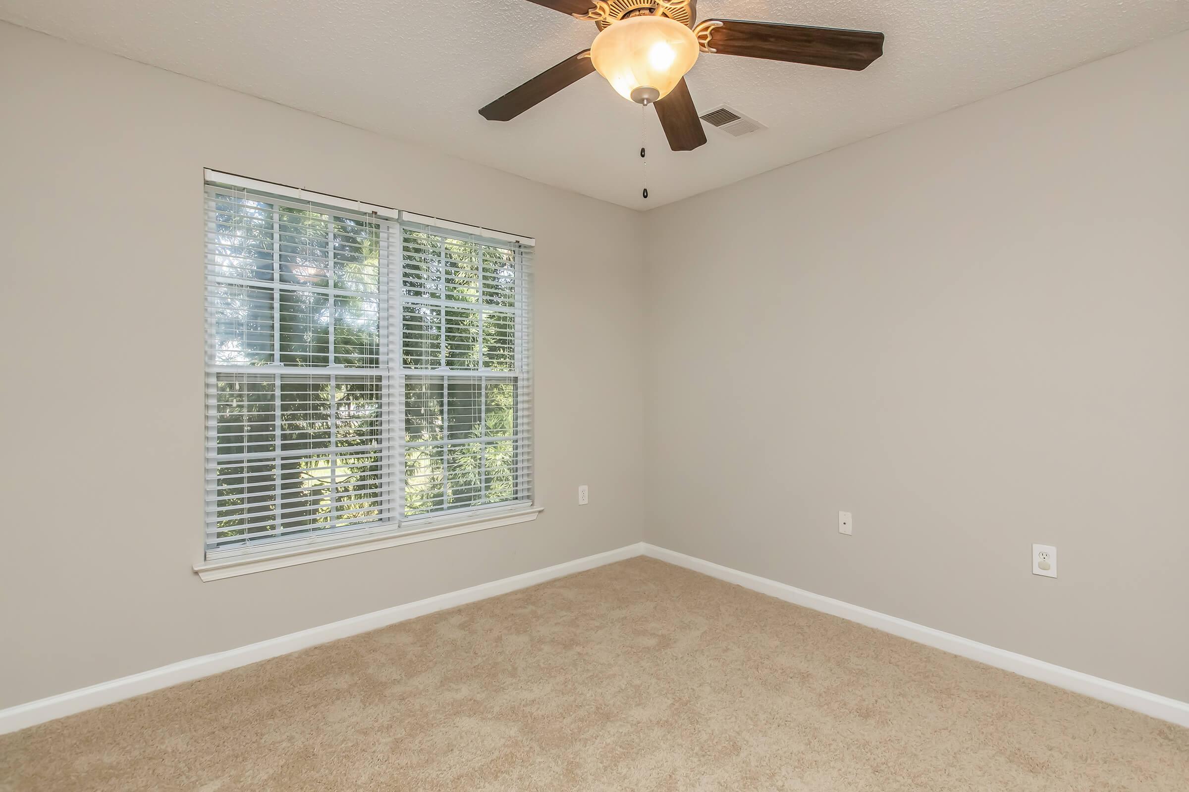 A well-lit room with beige carpet, a ceiling fan with light, and a large window featuring white blinds. The walls are painted a neutral color, and the space appears empty and airy.