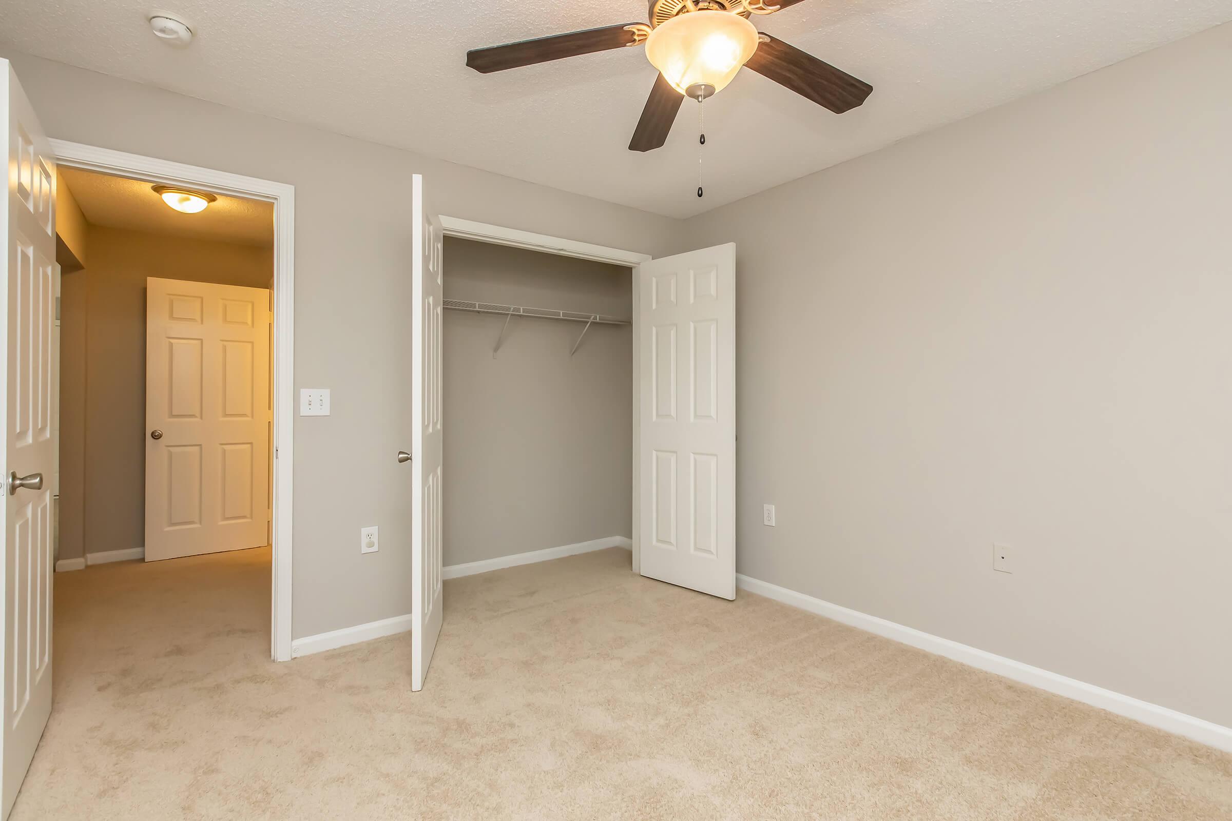 A well-lit, empty bedroom with light beige carpet and gray walls. There's a ceiling fan with wooden blades, and two open doors lead to a closet and another room. The space appears spacious and clean, with no furniture or decorations.