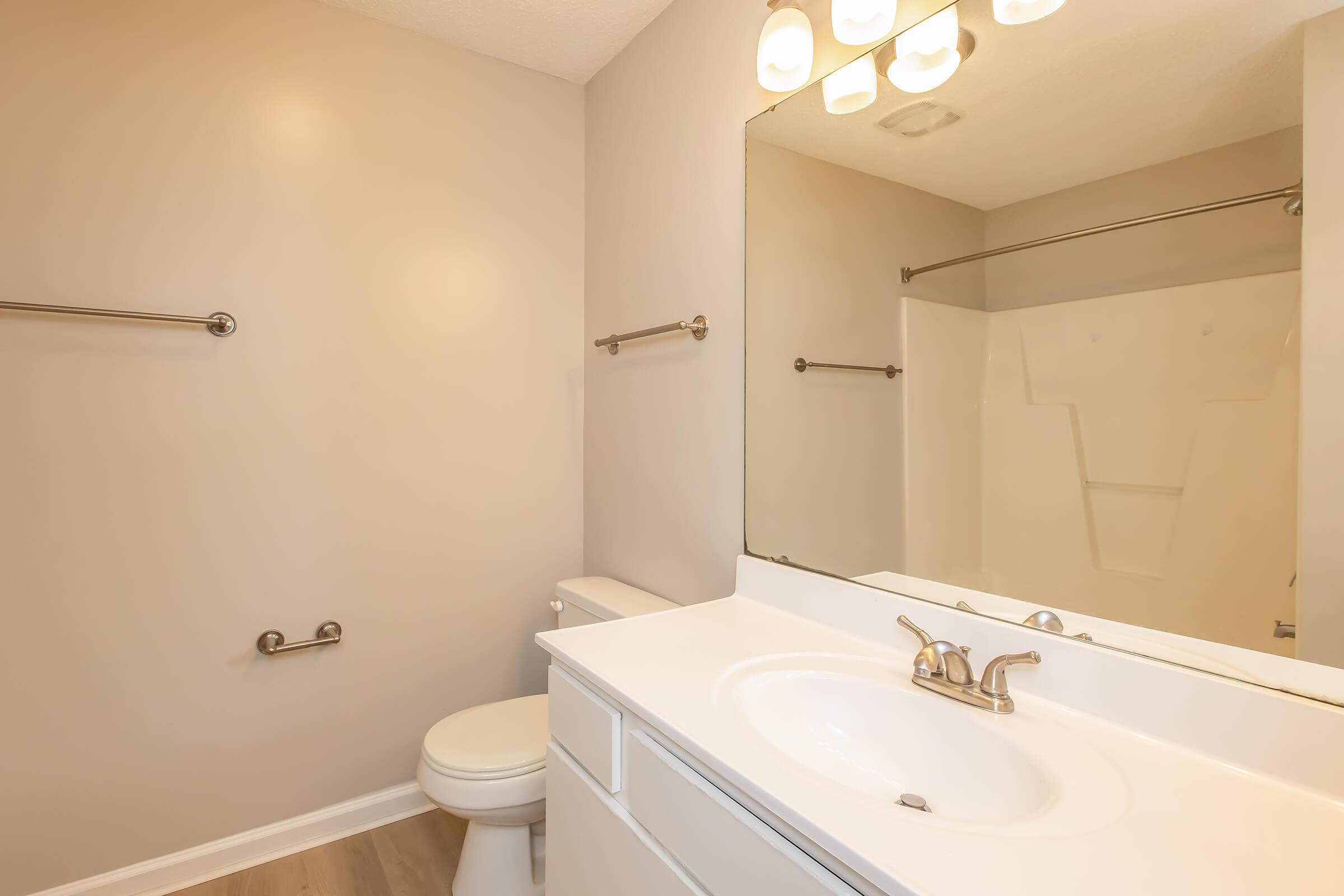 Bright and clean bathroom featuring a white vanity with a mirror, a toilet, a shower with a glass door, and towel racks. The walls are painted in a neutral beige, and the flooring is light-colored. Soft lighting from above adds warmth to the space.