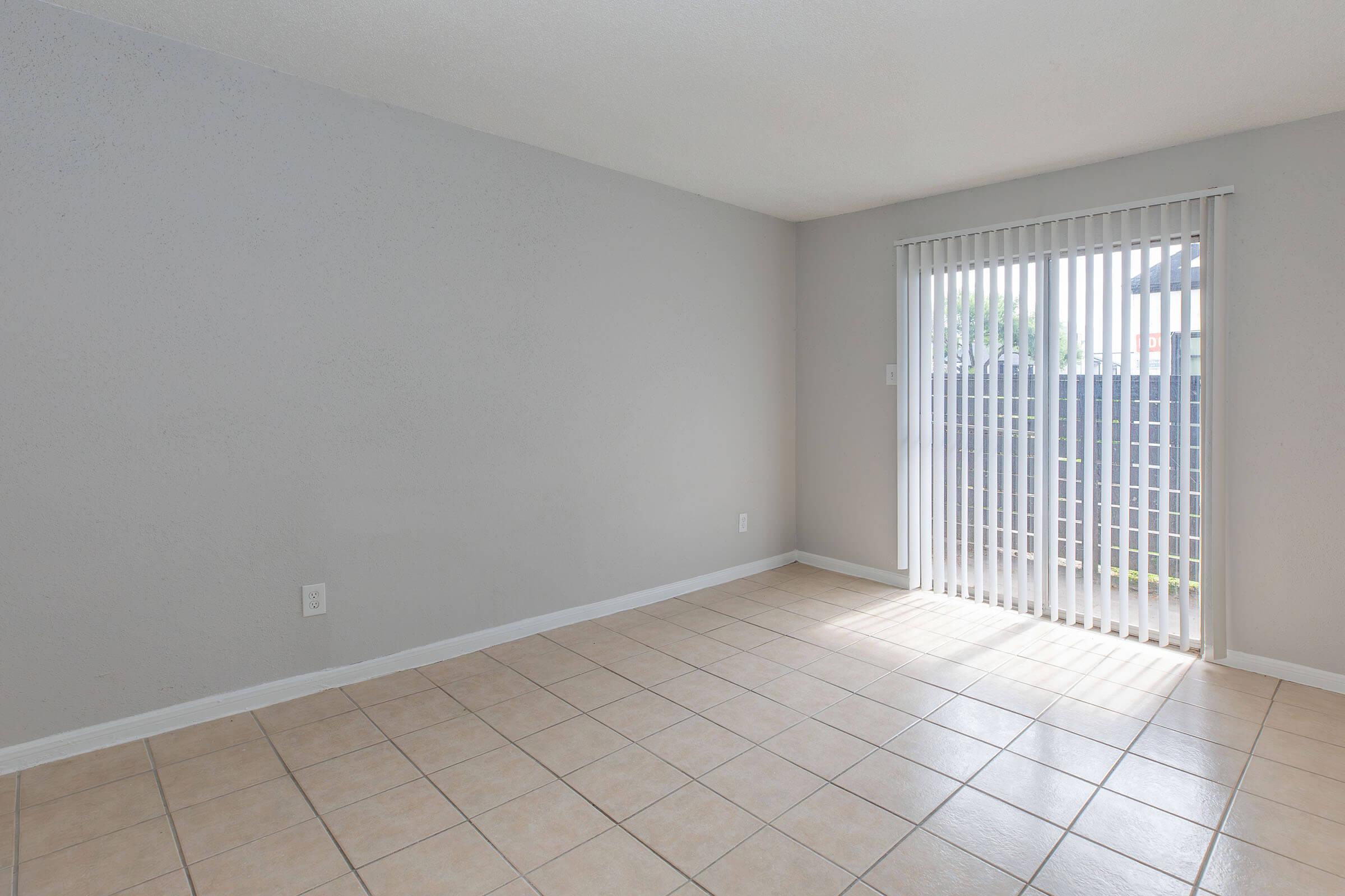 A vacant room with beige walls and tiled flooring. There are vertical blinds covering a large sliding door that leads to an outdoor area, allowing natural light to illuminate the space. The room is empty, giving a sense of openness and potential for various uses.