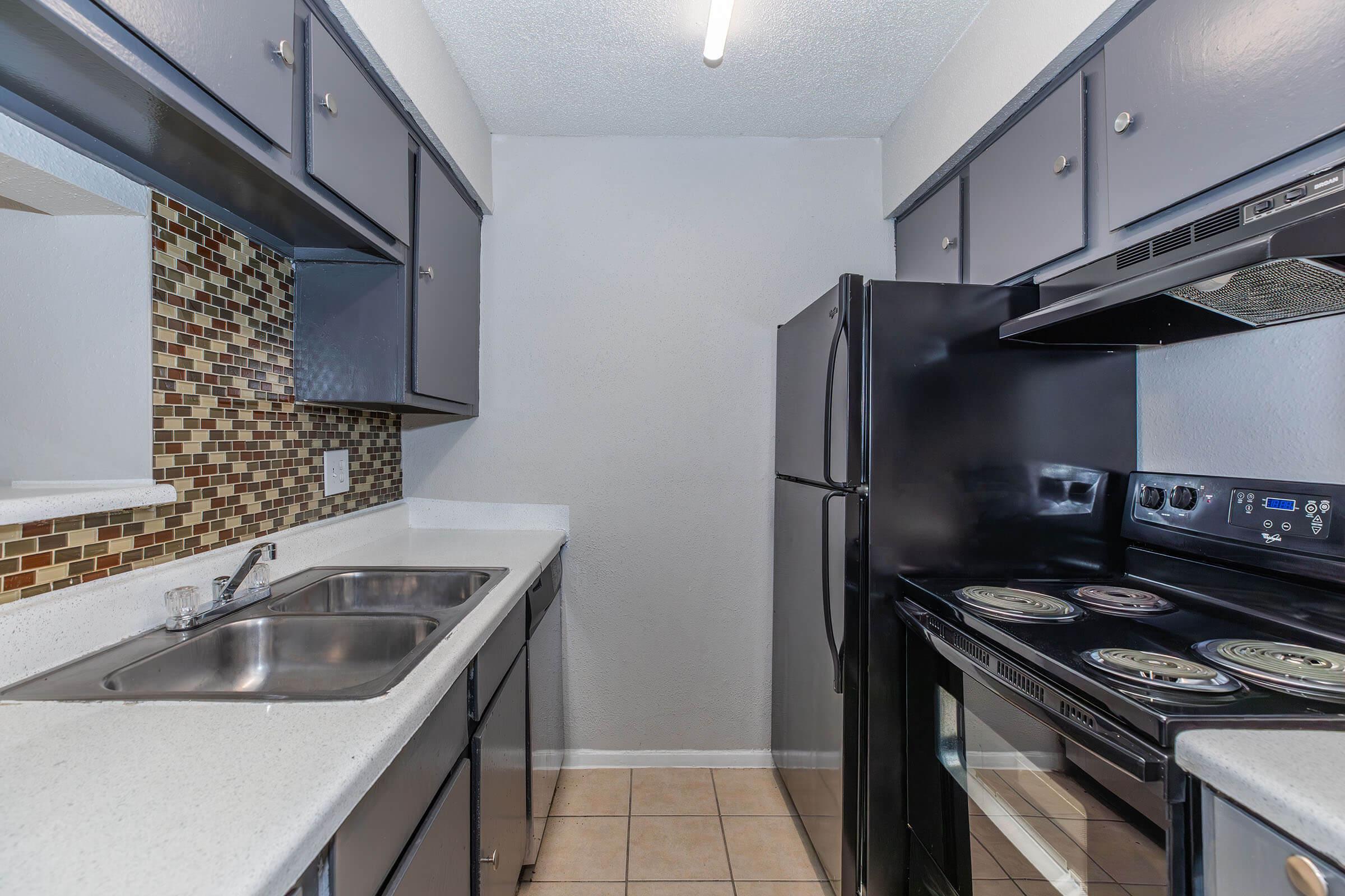 A modern kitchen featuring gray cabinets, a black refrigerator, and a black stove with an oven. The countertops are white, complemented by a mosaic backsplash. The space is well-lit with overhead lighting, and the floor is tiled with light-colored tiles.