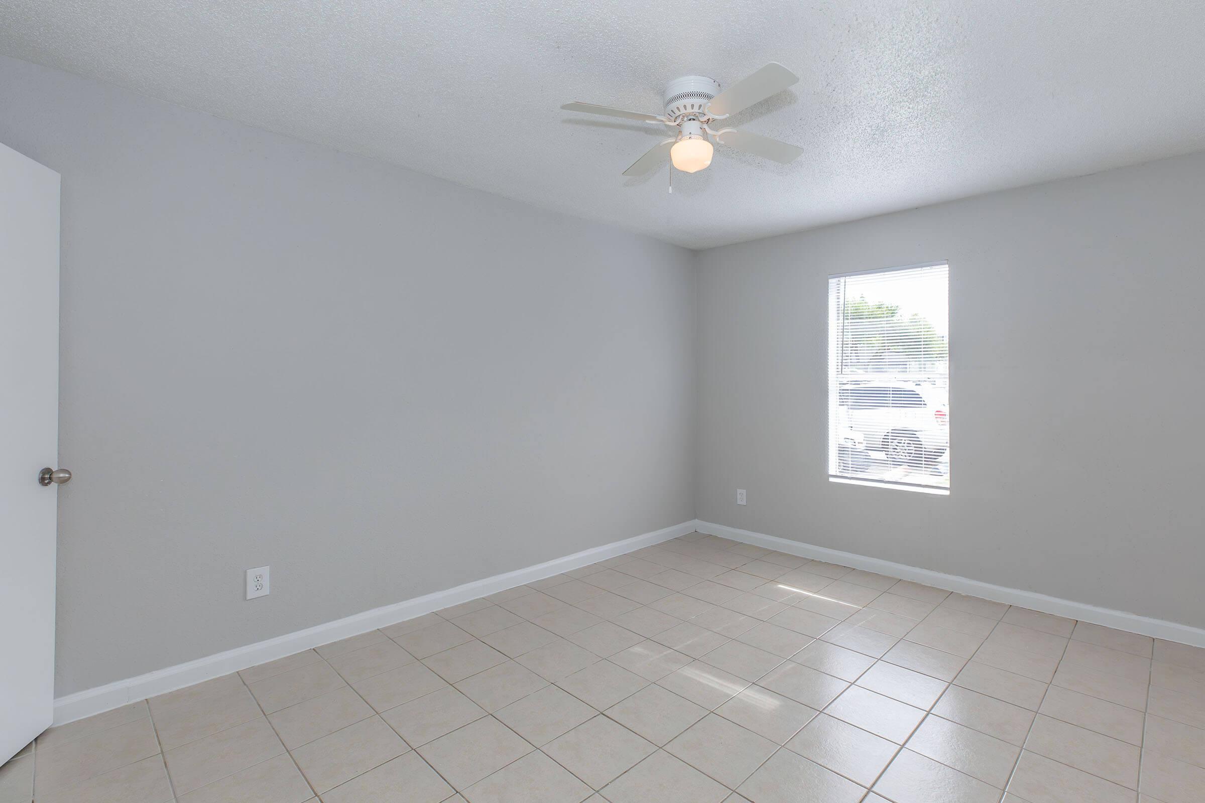 Empty room with light gray walls and a ceiling fan. The floor is tiled in a light color, and there is a window letting in natural light. A white door is visible, and the overall space appears clean and well-lit. The setting is unadorned, suggesting it could be used for various purposes.
