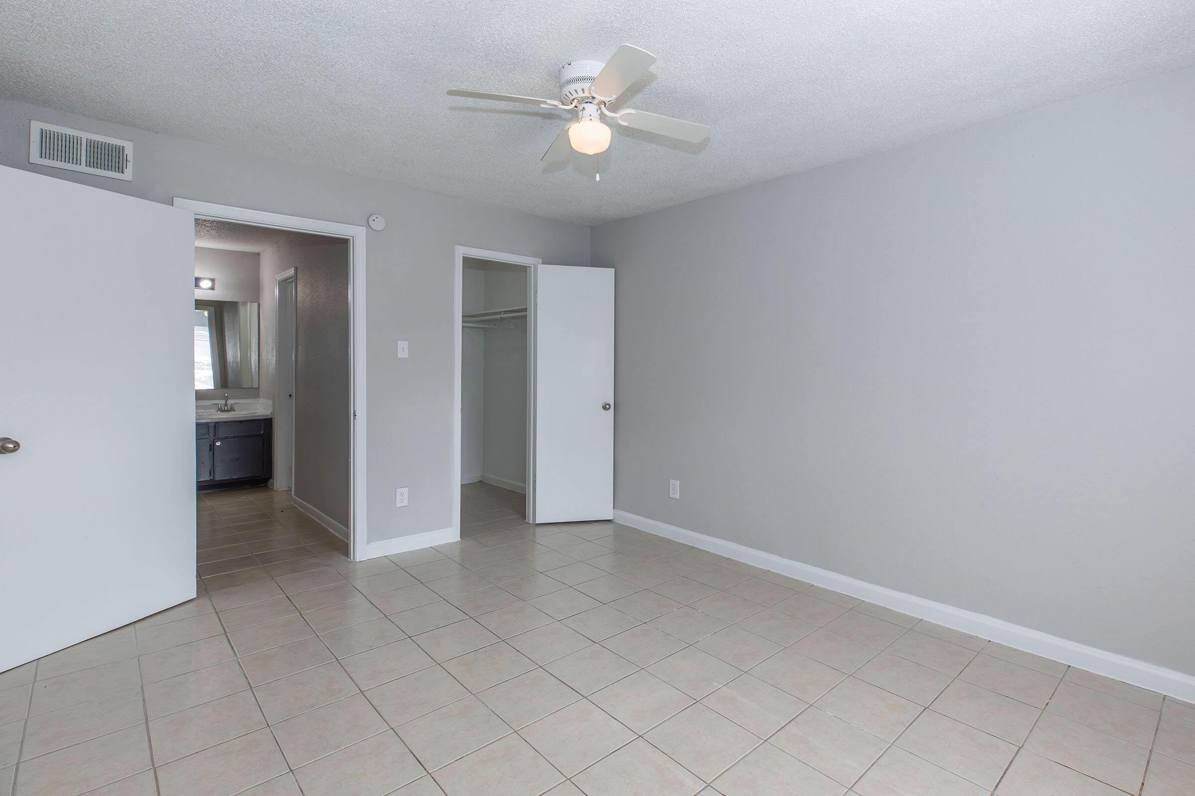 A spacious room with light gray walls and tiled flooring. A ceiling fan is overhead. Two white doors lead to a closet and a bathroom. Natural light is visible from the bathroom area, enhancing the bright and clean atmosphere of the space.