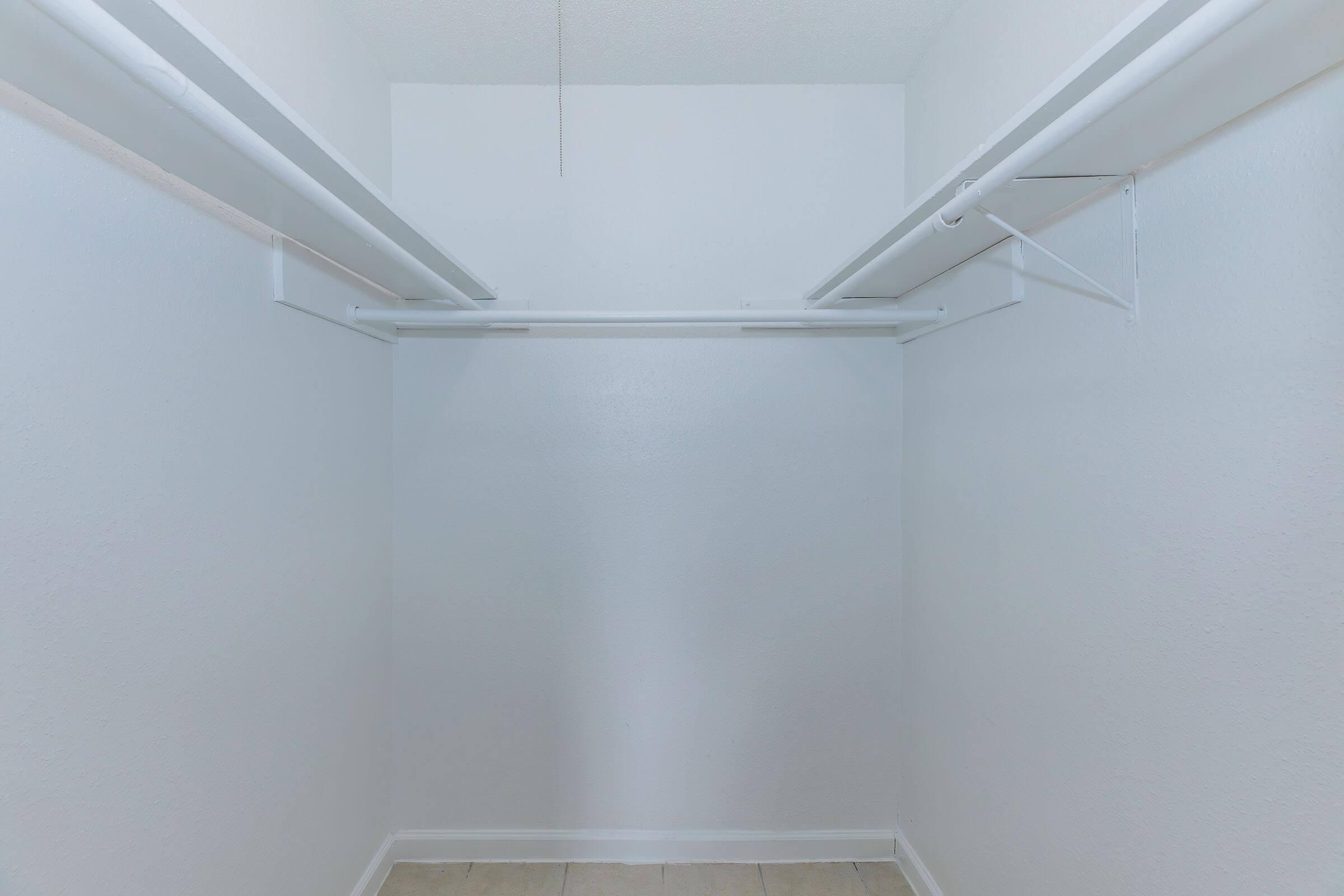 Empty closet with white walls and two horizontal hanging rods. The floor is tiled, and there is a light fixture visible on the ceiling. No items or furniture are present, creating a spacious and minimalist appearance.